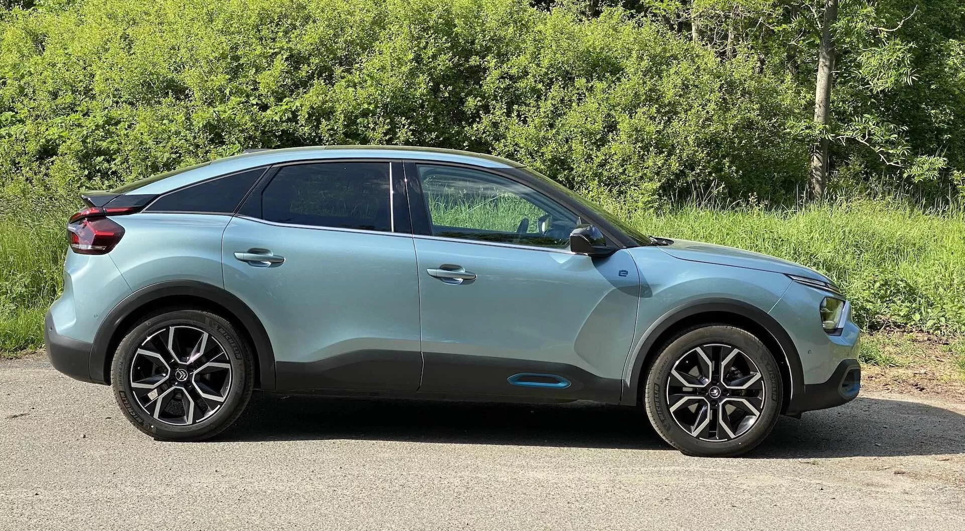 A 2020-as Citroen C4 (e-C4) oldalsó profilja, amely a vezető oldalát enyhén elölről szögben mutatja. A kép kiemeli a jármű modern formatervezési vonalait, jellegzetes többrészes kialakítású könnyűfém keréktárcsáit, karosszéria színű oldalsó tükreit integrált irányjelzőkkel, és a hátsó ablakok sötétített üvegét. A tető hátrafelé lejt, kupé stílust kölcsönözve a crossover/SUV karosszériának. Az autó fémes festékkel rendelkezik, amely kontúrokat és formatervezési vonalakat tartalmaz, lapos felületen parkol, háttérben zöld lombazattal, természetes napfény által megvilágítva.