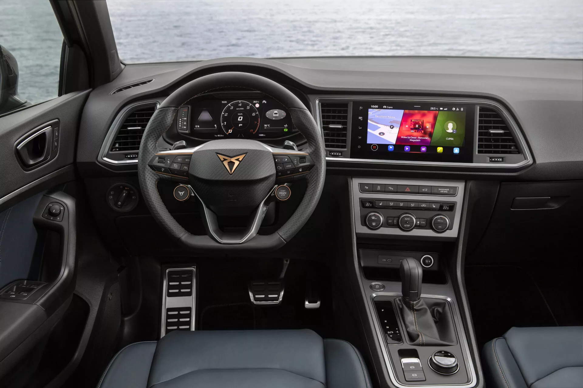 2020-as Cupra Ateca belső nézete a hátsó ülésekből, bemutatva a műszerfalat, a gyártó emblémájával ellátott multifunkcionális kormánykereket, a digitális műszerfalat, a vezető felé orientált infotainment érintőképernyőt, a klíma vezérlő gombokat és a modern váltógombot