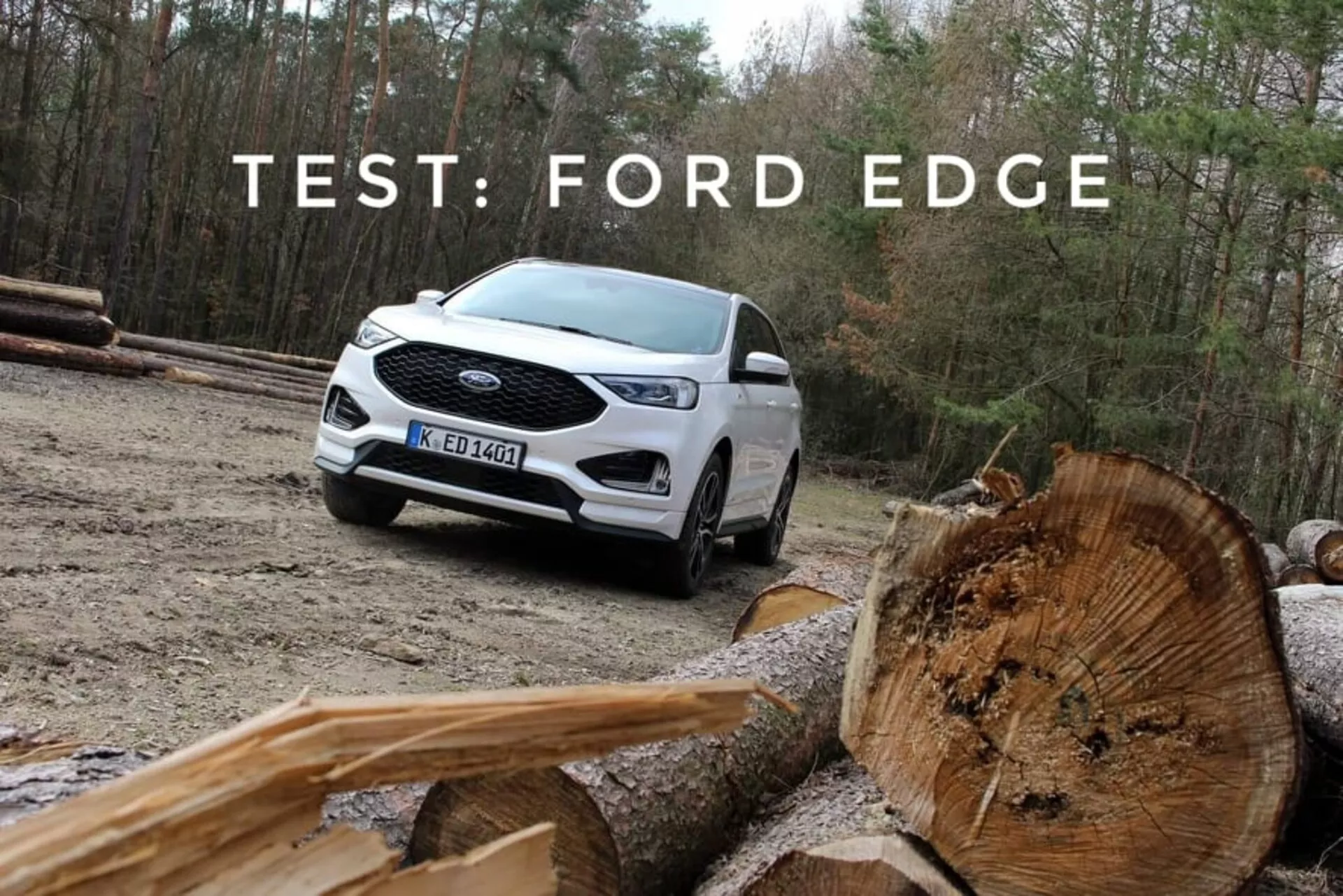 2018-as fehér Ford Edge, bemutatva terepjáró képességét egy háromnegyedes első és oldalnézettel, kiemelve a LED nappali menetfényeket, több küllős ötvözet keréktárcsákat, és kétszínű festést, természetes hátterrel és előtérben lévő vágott fa tuskókkal