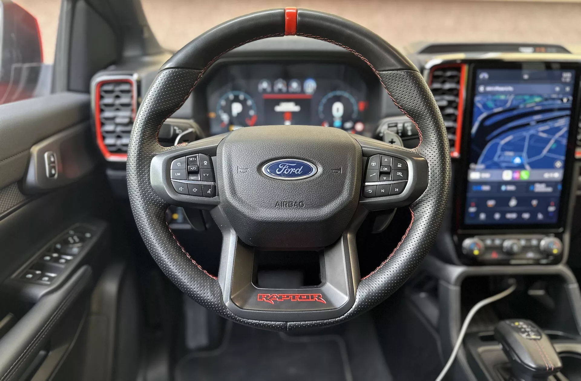 2022-es Ford Ranger Raptor belső előnézete, kiemelve a piros varrással ellátott, bőrbevonatú, háromküllős kormányt Ford logóval, a digitális műszerfalat, a modern érintőképernyős infotainment rendszert navigációs módban, a sportos műszerfalon lévő piros díszítéseket és a fejlett vezetői segédrendszereket a minőségi, technológia-orientált vezetési élmény érdekében