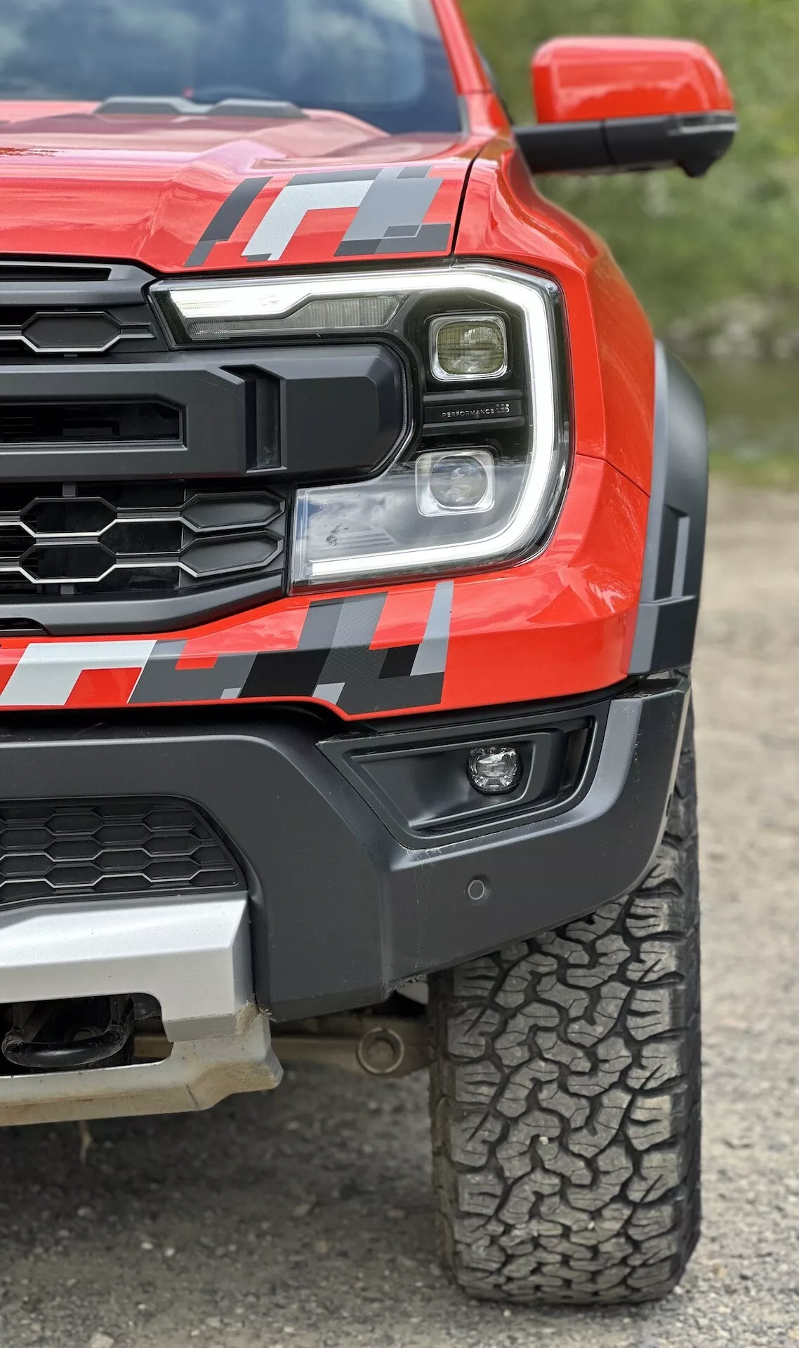 Közeli nézet egy 2022-es vörös Ford Ranger pickup-ra, bemutatva a LED fényszórót, erős hűtőrácsot, izmos első sárvédőt dekállal, és terepgumi mintázatot alacsony szögből, hogy kiemelje a robosztusságot és a fejlett technológiát