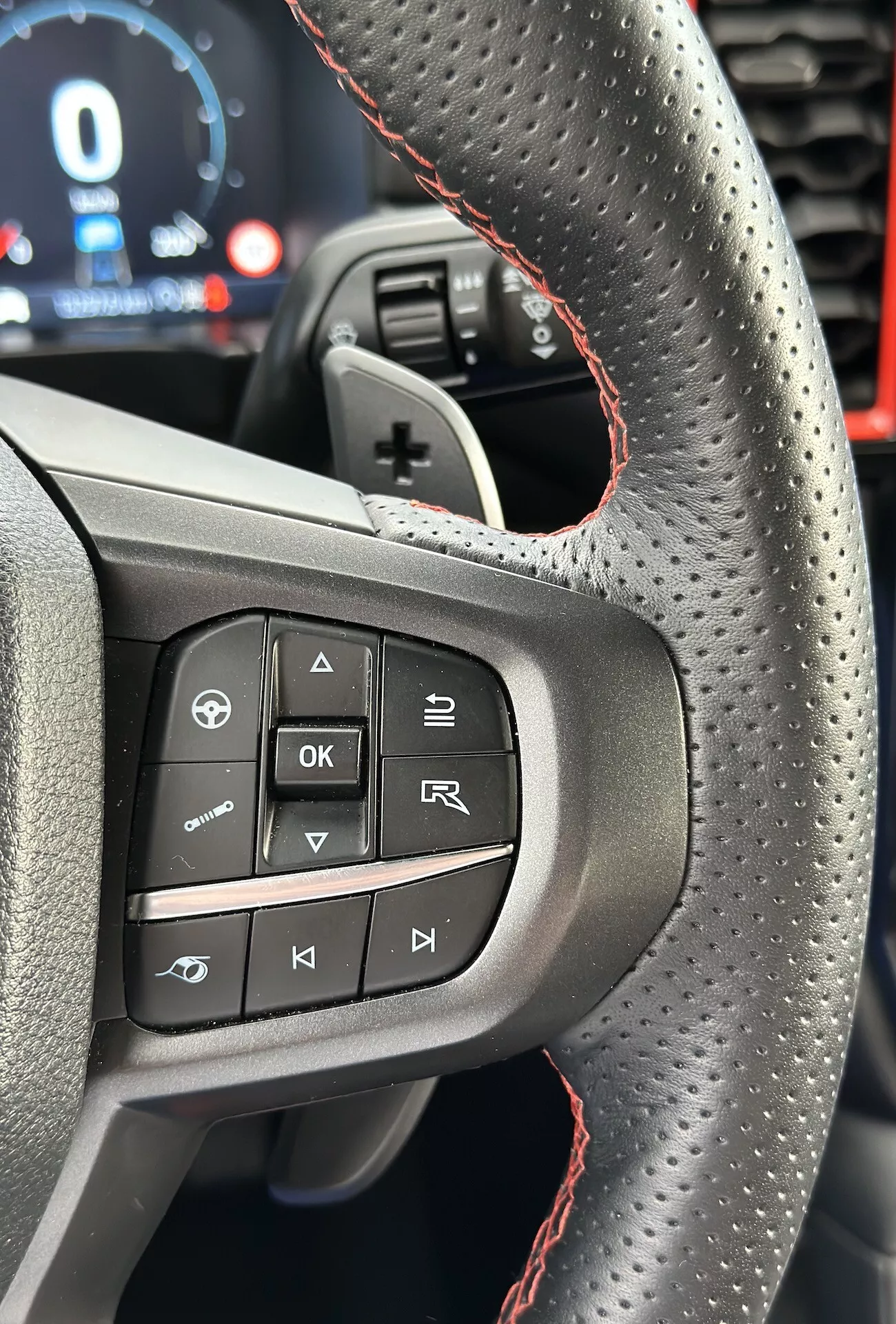 A 2022-es Ford Ranger kormánykerékének közelképe, kiemelve a bal oldali gombokat és jellemzőket, beleértve a lyukacsos bőrt piros varrással, a vezető perspektívájából, portré tájolásban