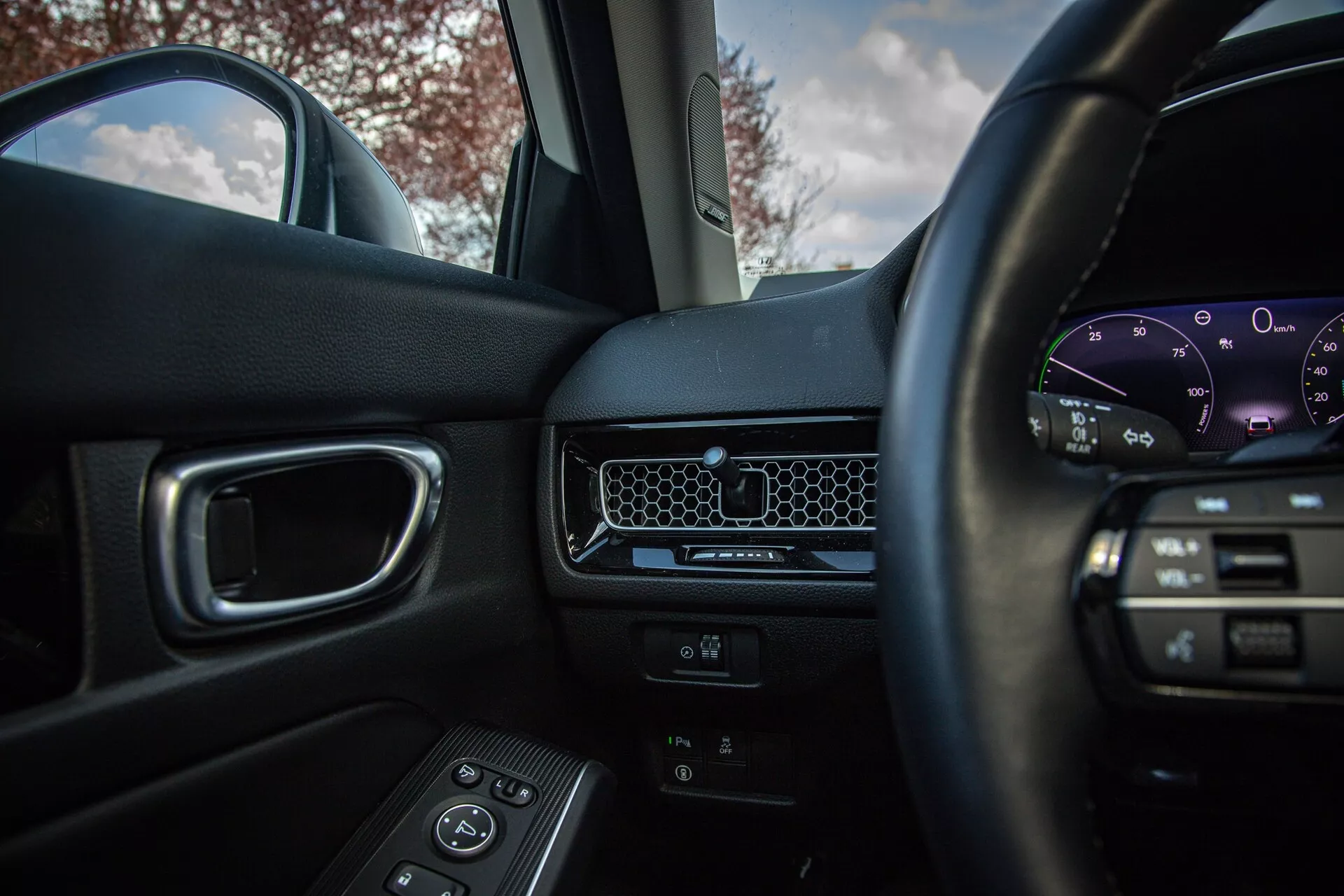 2022-es Honda Civic belső nézete a vezetőülésből, mutatva a médiavezérlőkkel ellátott kormányt, minimalista műszerfalat méhsejtes szellőzővel, digitális műszerfalat, utasajtót elektromos ablakvezérlőkkel és sötét színű első utasülést természetes fényben