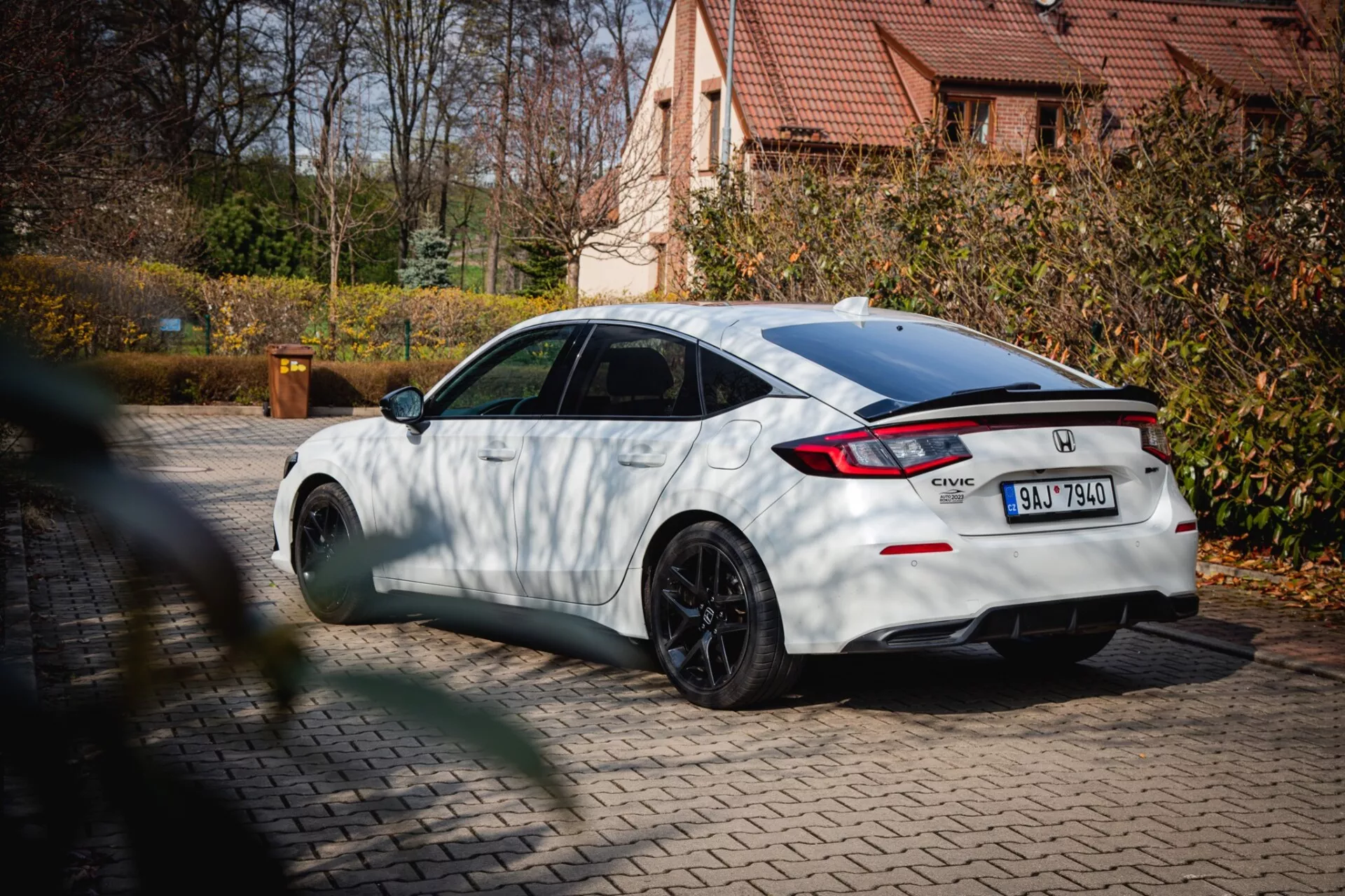 2022-es fehér Honda Civic fekete többsugarú kerekekkel és hátsó spoilerrel, parkolva egy járdán, őszi lombhullatással és lakóházzal a háttérben