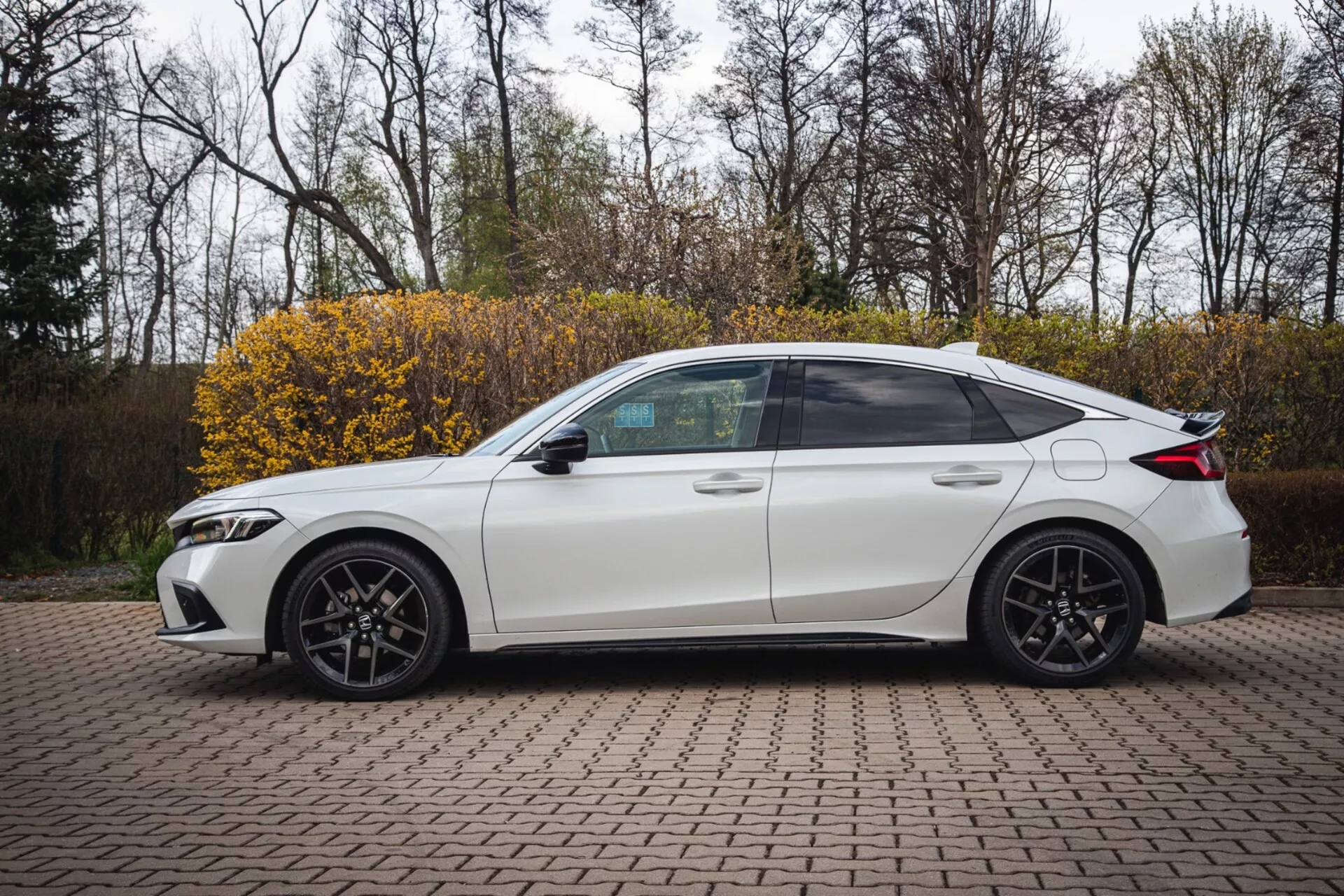 2022-es Honda Civic fehér négyajtós szedán, parkolva egy térkövezett területen, oldalnézeti profilban sportos tervezéssel és modern LED fényszórókkal, kissé emelt szögből fényképezve az oldal teljes nézetének kiemelése érdekében, beleértve a sötétített ablakokat, sportos ötvözetű kerekeket és technológiai jellemzőket, mint például az oldalsó tükrökbe integrált irányjelzők.