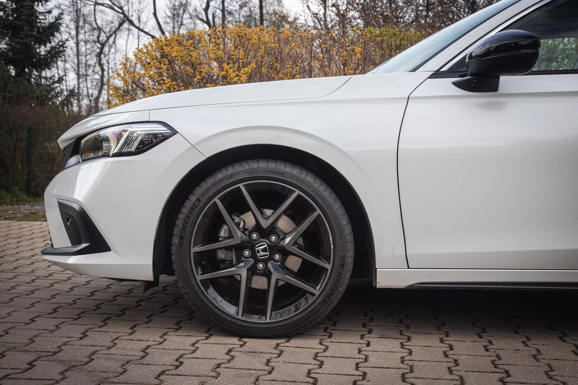 2022-es fehér Honda Civic, amely bemutatja a bal első kereket többsugaras ötvözött kialakítással és alacsony profilú abronccsal, valamint a sárvédő részleteit és az első lökhárító részét téglaburkolaton