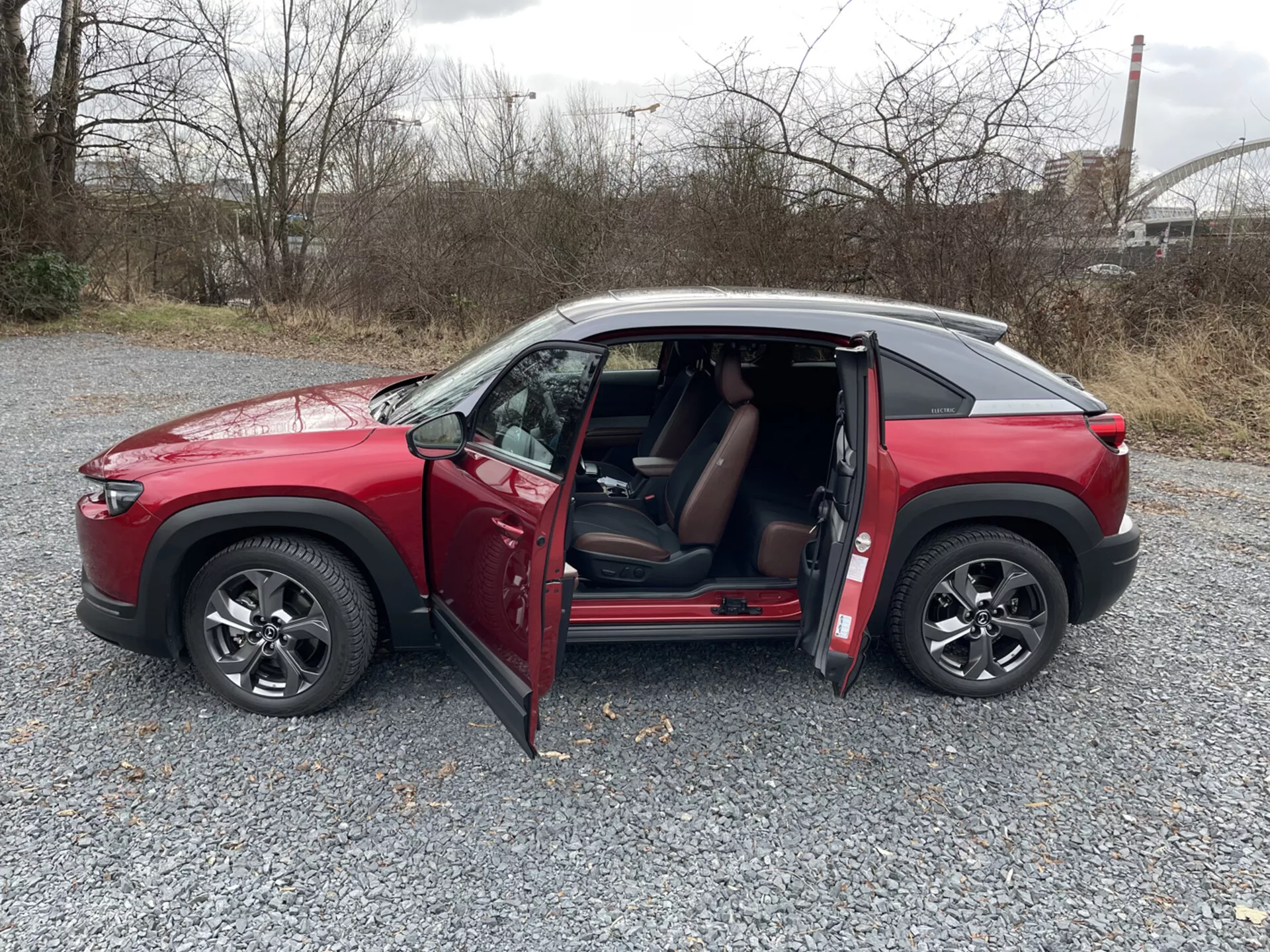 Piros, négyajtós Mazda MX-30 SUV 2020-es modell profilnézetben, nyitott ajtókkal, többsugaras ötvözött kerekekkel, sötétített ablakokkal és kontrasztos színű tetővel ipari környezetben és zöldterülettel a háttérben.