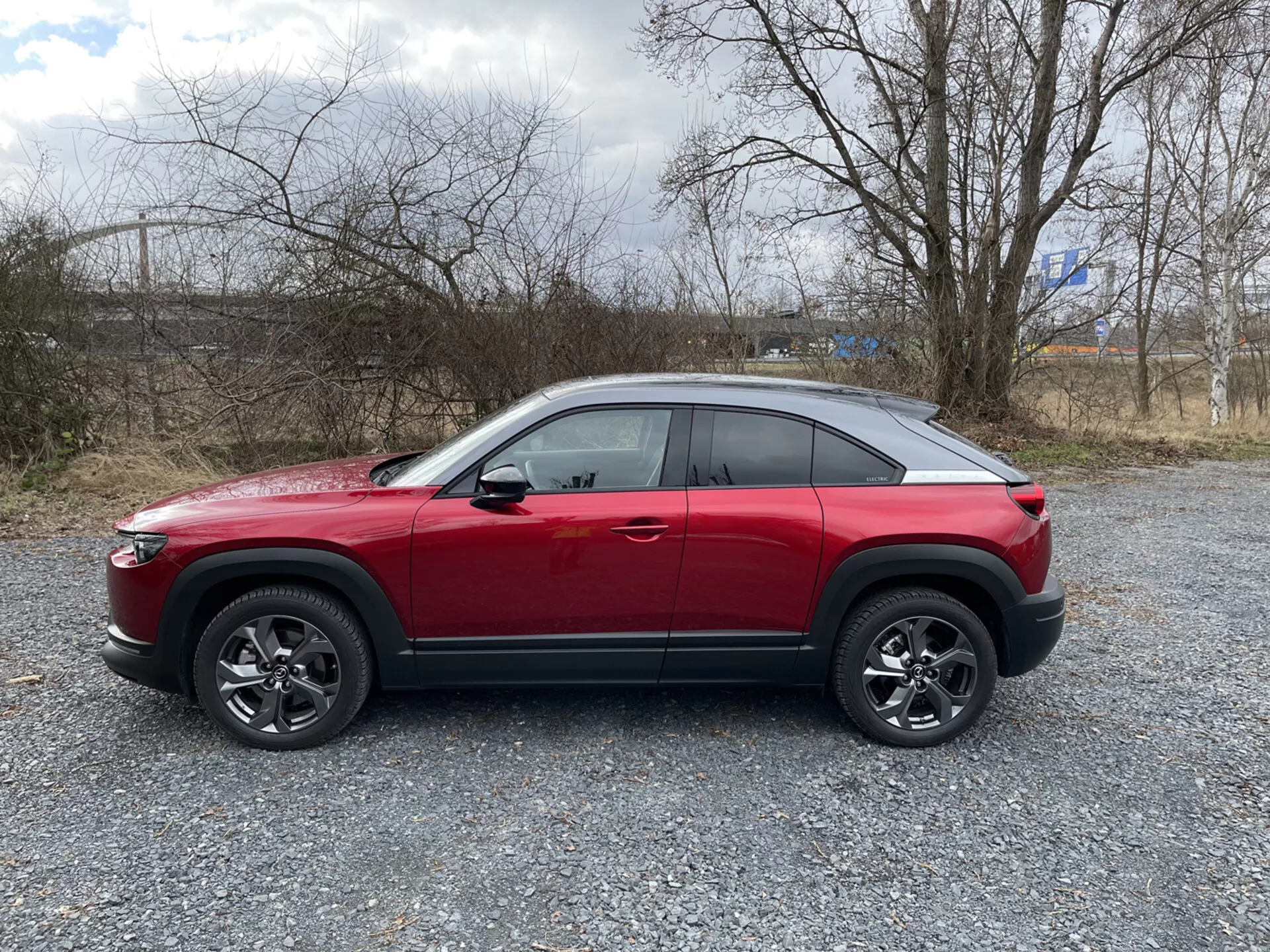 2020-as Mazda MX-30 piros crossover SUV oldalnézete sóderes talajon, ritkás növényzettel és felhős éggel a háttérben, bemutatva magas övvonalat, kupé-szerű lejtős tetővonalat, dinamikus karaktervonalat, többspókás ötvözött kerekekkel fekete műanyag burkolattal, és lehetséges fejlett technológiai jellemzőket, úgy mint ADAS, LED nappali menetfény, kulcs nélküli belépés, TPMS és infotainment rendszer.