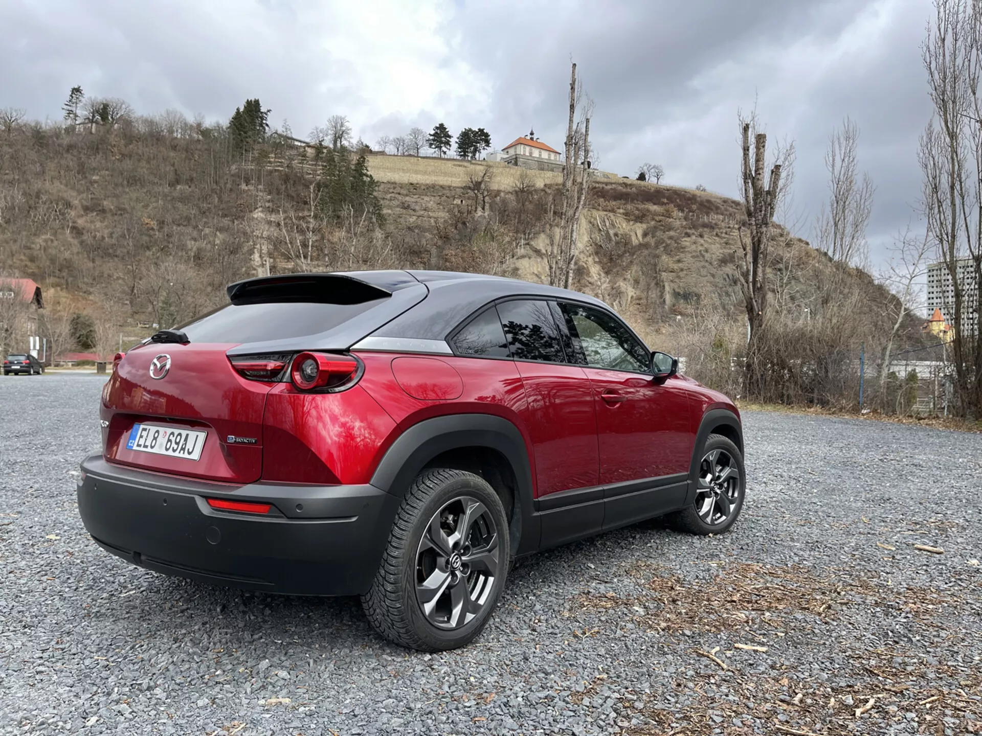 2020-as Mazda MX-30 modern crossover parkol a sóderen, bemutatva sportos, piros-fekete kéttonusú tervezését, LED hátsó lámpáit, hátsó légterelőjét és többsugaras ötvözött kerekeit természet és építészet háttérrel