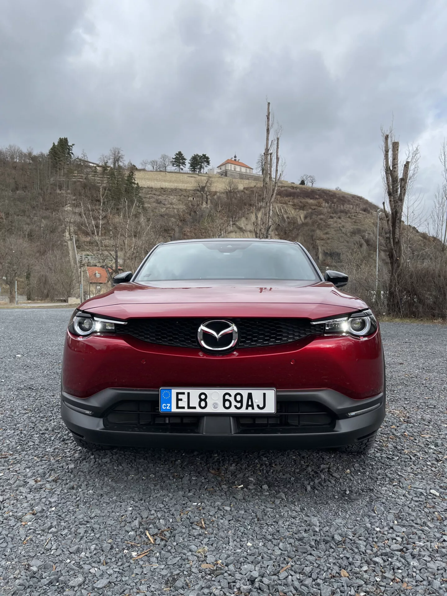 Egy 2020-as évjáratú, piros Mazda MX-30 eleje, kiemelve az autó hűtőrácsát, lökhárítóját és LED fényszóróit, kavicsos talajon, felhős égbolt, zöldellő táj és egy épület háttérrel