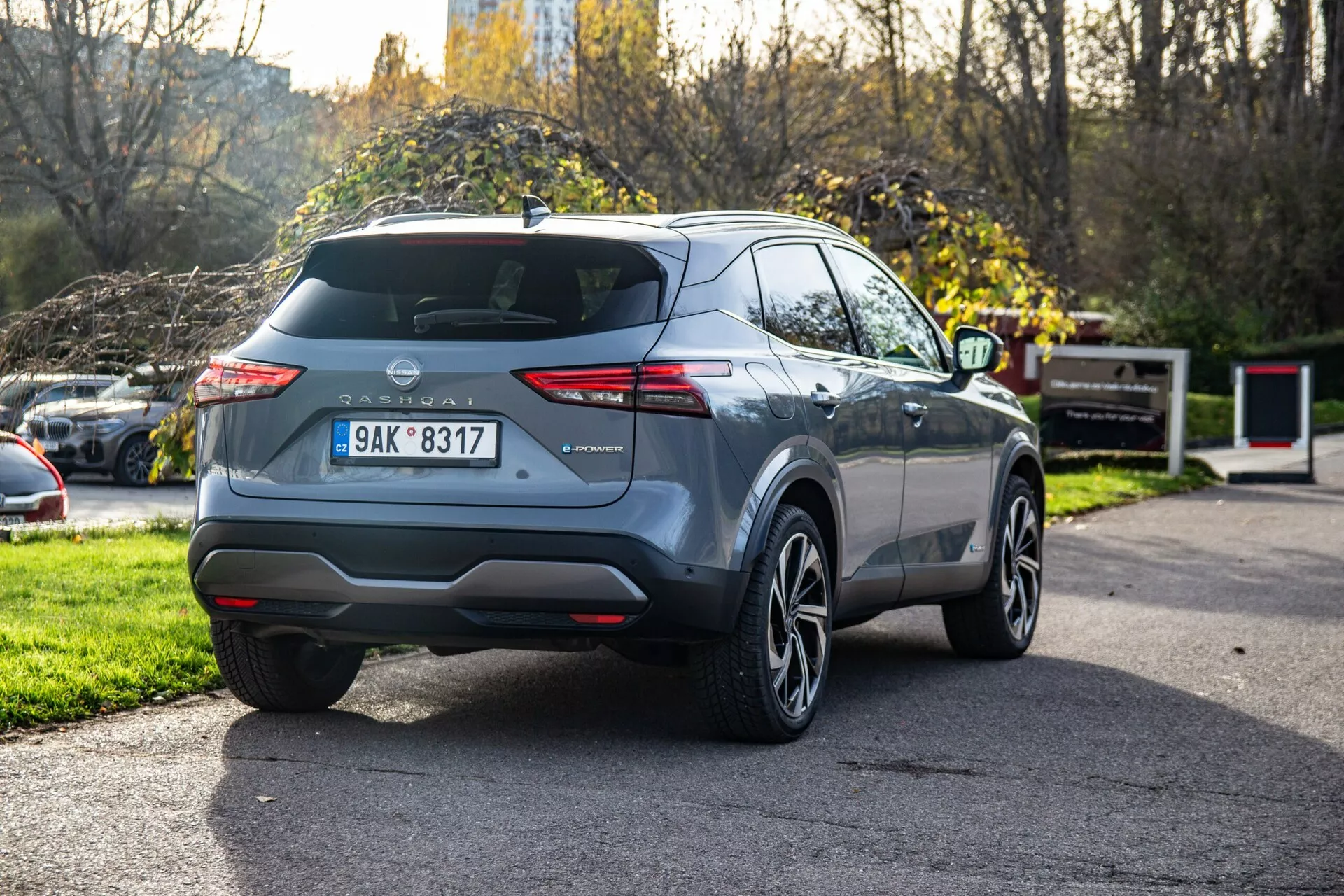 2021-es Nissan Qashqai jármű hátsó háromnegyedes szögből fényképezve, kiemelve a jellegzetes hátsó lámpatesteket, a látható hátsó ajtót ablaktörlővel, középen elhelyezett gyártói emblemát, középen a hátsó lökhárítón elhelyezett rendszámtáblát, integrált hátsó lökhárító reflektorokat, egyetlen kipufogóvéget, könnyűfém kerekeket, modern karosszéria vonalakat, keskenyedő oldalablakokat, lehetséges tetősíneket, rövid tetőantennát, fémes festésű kivitelét és részben látható oldalsó tükröt.