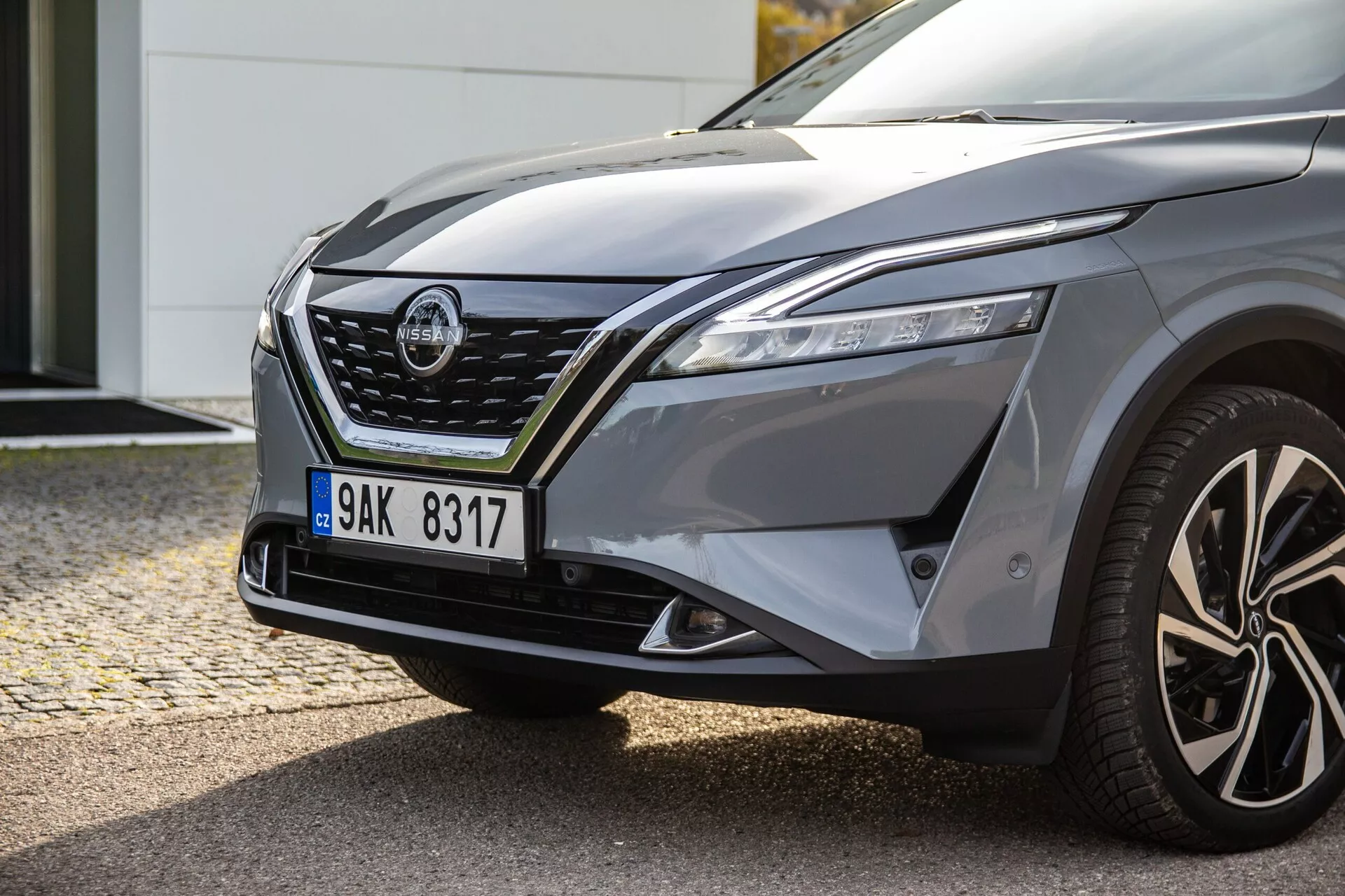 Modern autó bal első oldalának közelképe, amely elegáns LED fényszórókat, kiemelkedő krómozott rácsokat, formázott motorháztetőt, domború sárvédőt, többrudas könnyűfém kerekeket és integrált irányjelzőkkel ellátott vezető oldali tükröt mutat, tükrözve az autó dinamikus kialakítását és potenciális kezelési képességeit.