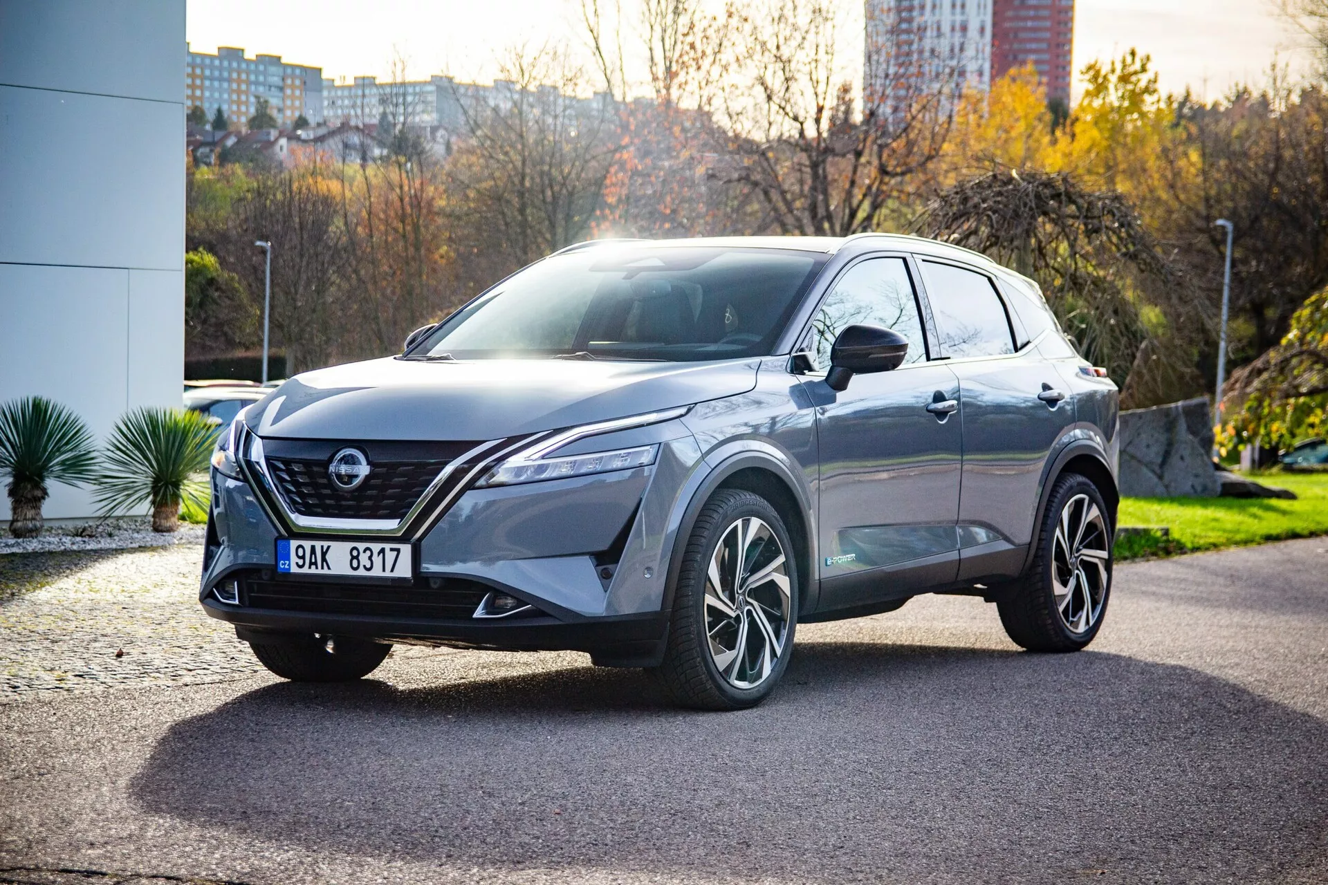 Háromnegyedes elülső nézet egy modern Nissan Qashqai 2021 crossover SUV-ról, ferdén elhelyezve a képkockán egy burkolt területen, városi háttérrel növényzettel és épületekkel. A látható fő jellemzők közé tartoznak a keskeny, szögletes LED fényszórók, egy kiemelkedő V-alakú hűtőrács, integrált ködlámpák, parkolószenzorok, formázott motorháztető, kifejezett kerékjárati ívek, többrudas ötvözet kerekek, az előrehaladott vezetést segítő rendszerekhez kapcsolódó érzékelő komponensek a hűtőrácson, karosszéria színű visszapillantó tükrök esetlegesen integrált irányjelzőkkel, panorámatető, sötétített ablakok, amelyek bőrrel bevont üléseket sugallnak, valamint aerodinamikus karosszériavonalak.