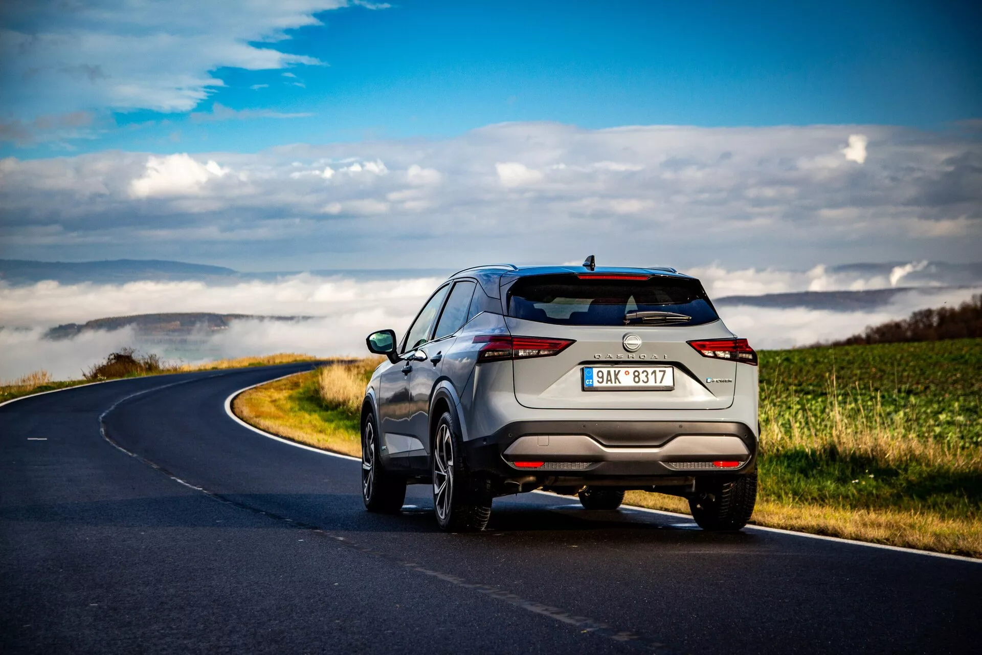 2021-es Nissan Qashqai SUV hátsó háromnegyed szögből ábrázolva egy kanyargós vidéki úton szétszórt felhők alatt, modern és elegáns formatervezéssel, vízszintesen elhelyezett keskeny hátsó lámpákkal, diszkrét kipufogókkal, többrugós könnyűfém kerekekkel, tetősínekkel és fejlett parkolószenzorokkal. A világos fémes szín kiemeli a prémium érzését.