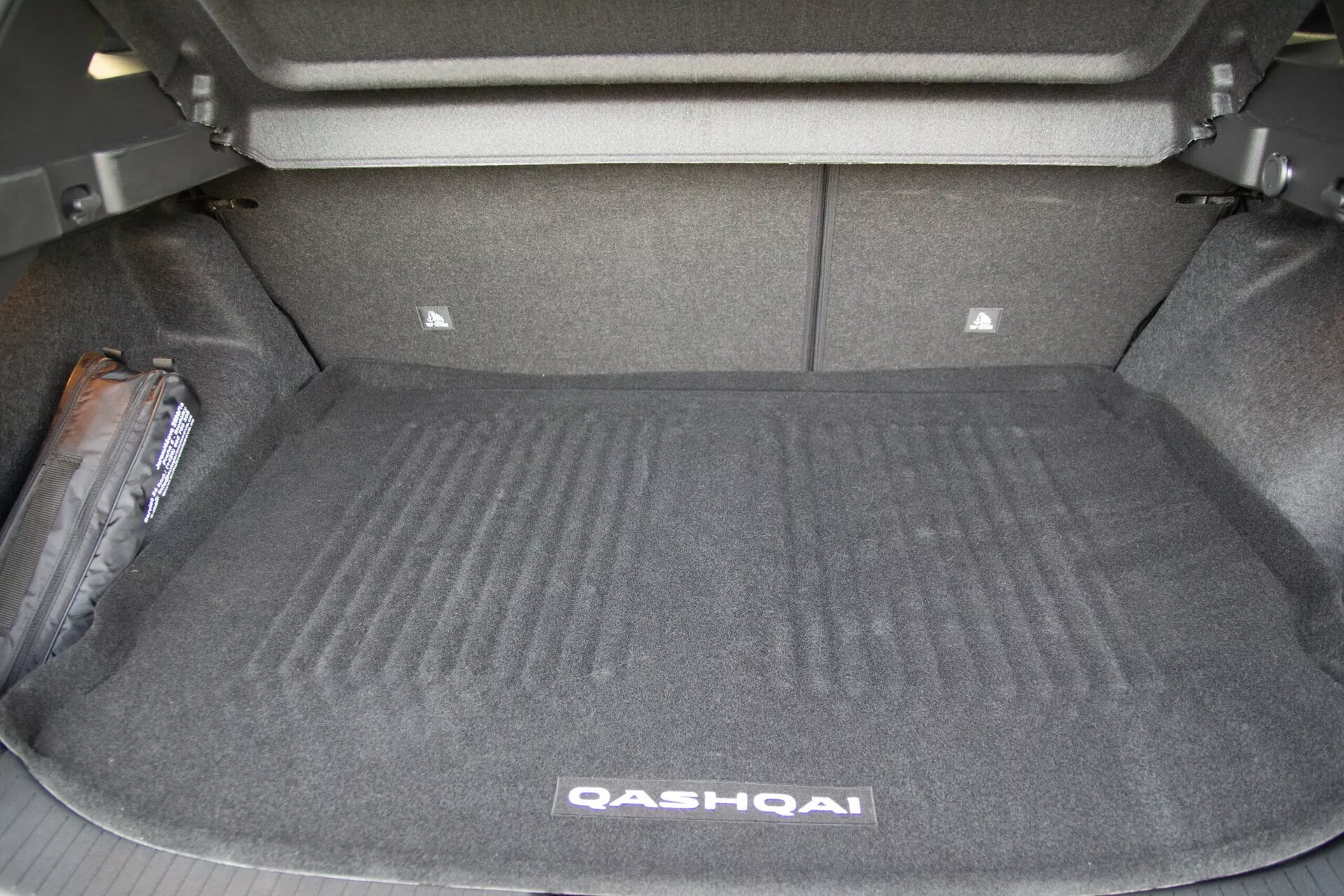 Kép, amely egy 2021-es Nissan Qashqai nyitott csomagterét mutatja hátulról. A rakomány nélküli csomagtér szőnyeggel borított felületét, oldalfalait és a függőleges helyzetben lévő hátsó üléseket felfedi. A csomagtér elején kissé megemelkedett terület az alsó gépészeti elemeket vagy a hátsó ülések megtámasztását javasolja. A csomagtér hátulján egy kisebb tároló rekesz látható. A bal oldalon van egy vészhelyzeti felszerelési cikk, valószínűleg egy emelő vagy elsősegély-készlet. A kép kiemeli a jármű tágas csomagtér kapacitását és funkcionális csomagtér tervezését.