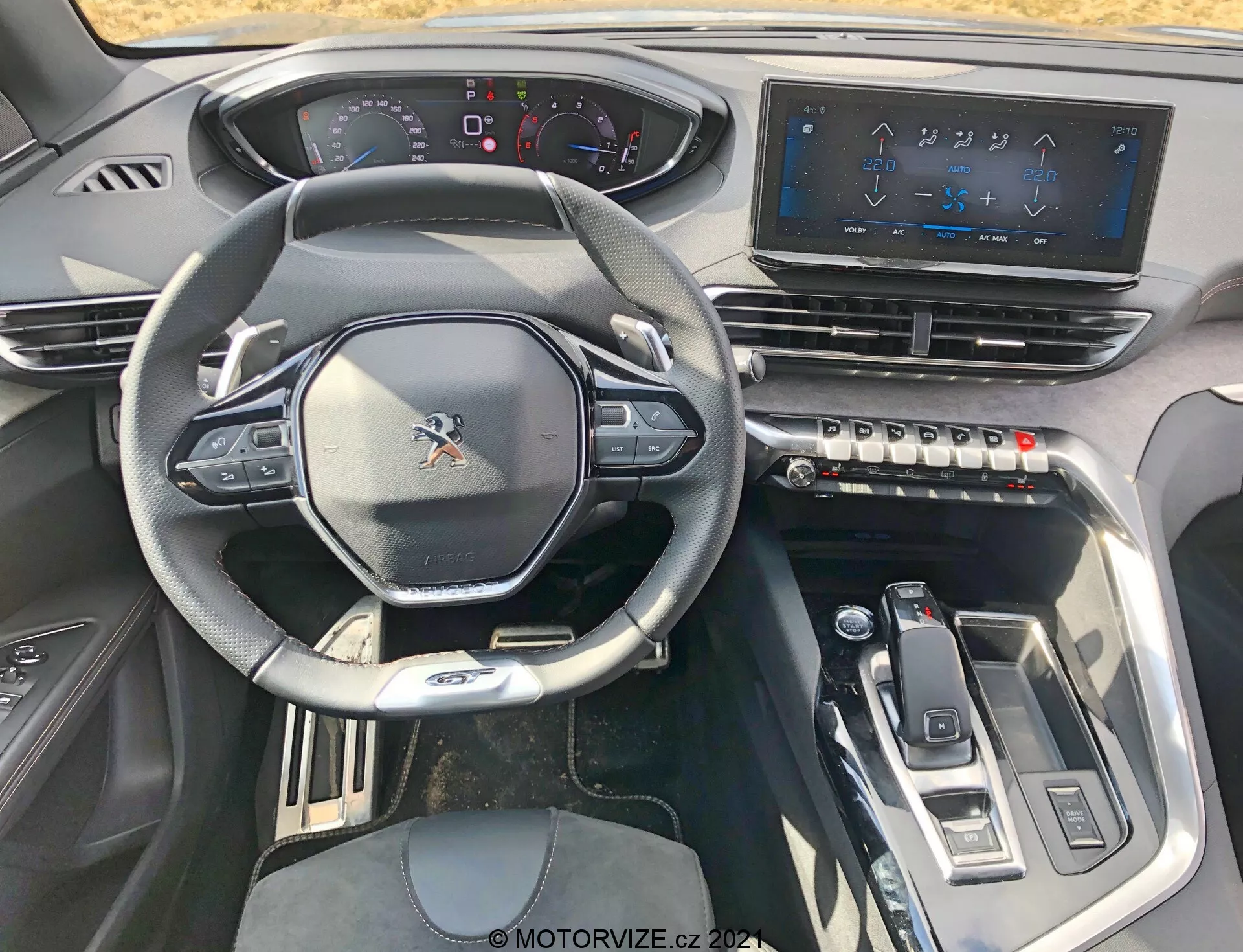 2021-es Peugeot 5008 belső nézete a vezetőülésből, sportos többfunkciós kormánykerékkel és beépített vezérlőkkel, részben digitális műszerfallal, nagy érintőképernyős infotainment rendszerrel, érintésérzékeny klímavezérlő gombokkal, elegáns váltóval és modern dizájnelemekkel, amelyek fémes díszítéssel magas szintű technológiai élményt nyújtanak.