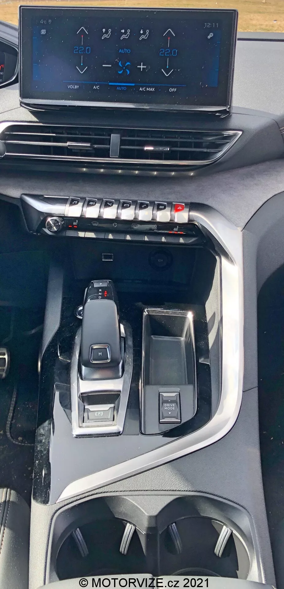 A Peugeot 5008 2021-es belső nézete a középkonzolra fókuszálva felülnézetből. A kép kiemeli a lebegő infotainment érintőképernyőt, amely a légkondicionáló vezérlőit mutatja, a fizikai gombok és tekerők fölött, amelyek a klímakontroll gyors beállításait szolgálják. Alatta egy elegáns, valószínűleg elektronikus sebességváltó kar látható, amelyet vezetési mód választó és egy multifunkcionális vezérlőgomb vesz körül. A tervezés egy modern, prémium járműre utal fejlett technológiai és ergonomikus funkciókkal, beleértve a pohártartókat és tárolóhelyeket.