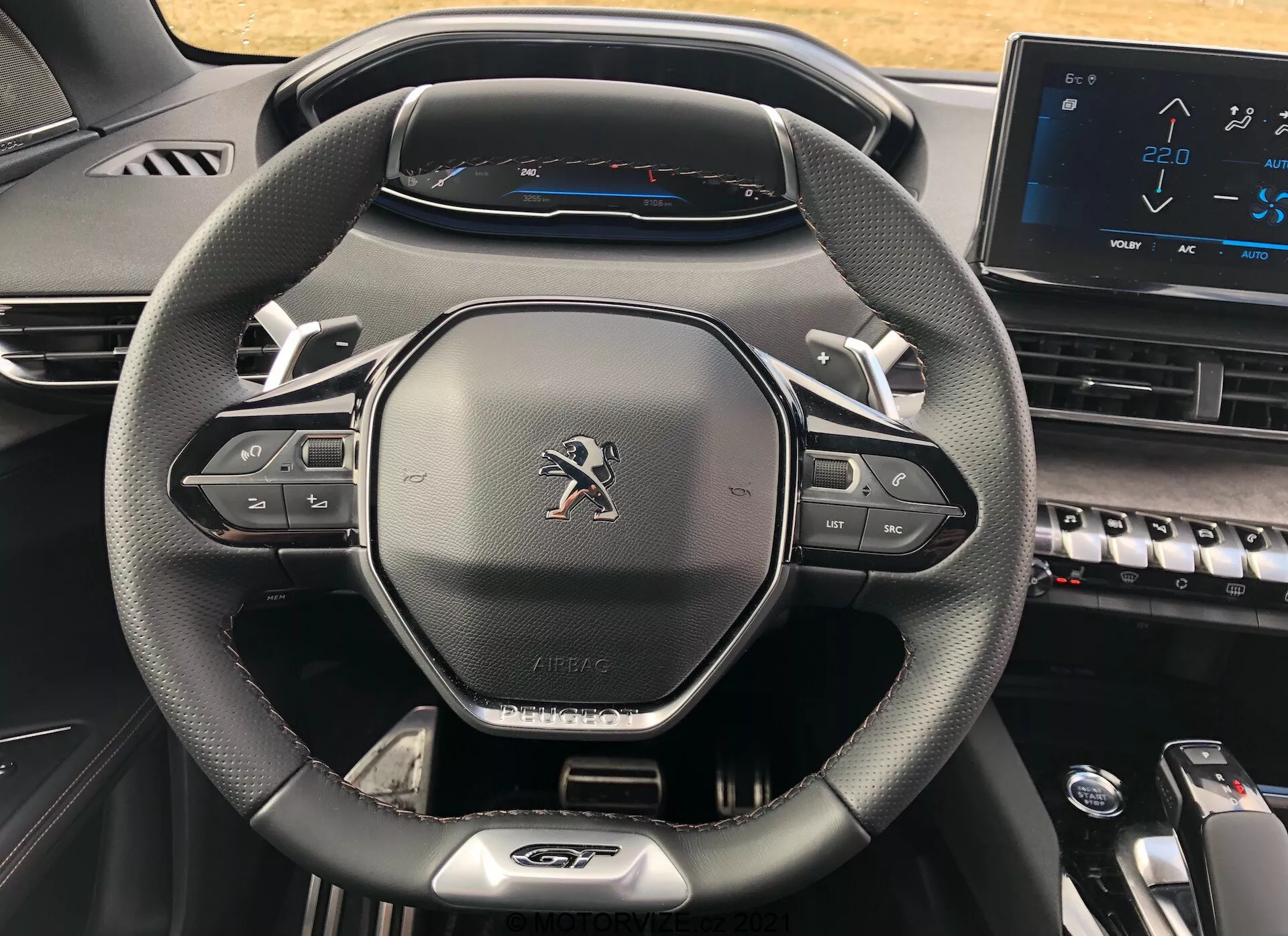 Közeli kép egy 2021-es Peugeot 5008 vezetői műszerfaláról, amely kiemeli a modern, háromküllős, bőrszerű anyagból készült kormánykereket multimédia és tempomat vezérlőgombokkal, Peugeot emblémával, 'AIRBAG' felirattal, teljesen digitális műszercsoporttal, amely 220 km/h végsebességet mutat, és a középkonzol részlegesen látható, pianó stílusú gombokkal és elektronikus érintőképernyővel, amely egy korszerű és technológiailag fejlett kialakítást mutat be.