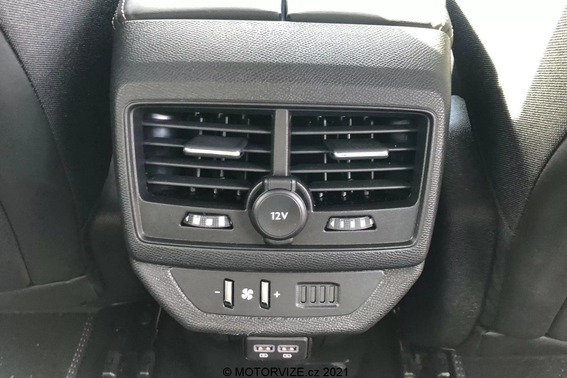 Közeli kép a Peugeot 5008 2021-es modell hátsó utasok számára kialakított funkcióiról a középkonzol hátoldalán. A képen két légbeömlő, egy 12V-os csatlakozó, ülésfűtés vezérlők, USB portok és egy üres gomb látható, hátsó ülésen ülő szemszögéből fényképezve.