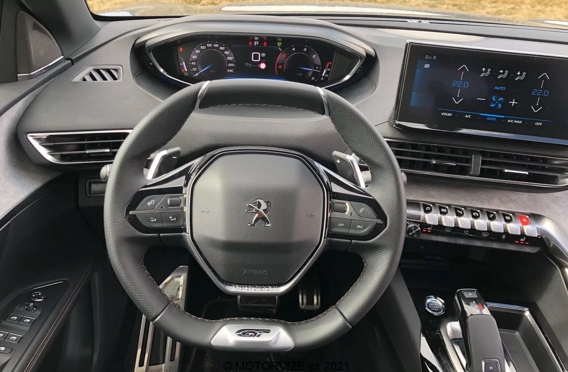 Peugeot 5008 2021 belső nézete a vezetőülésből, kiemelve a bőrborítású, lapos alsó kormánykereket, amelyre szerelt kezelőszervek vannak, a teljesen digitális műszerfal világító kijelzőkkel, a középső konzolon lévő nagyméretű érintőképernyős infotainment rendszert, elegáns légkondicionáló vezérlőket, elektronikus sebességváltó választót az automata sebességváltóhoz és a prémium, sötét anyagokat tartalmazó dizájnt, amely modern, csúcstechnológiai és prémium esztétikát sugall.