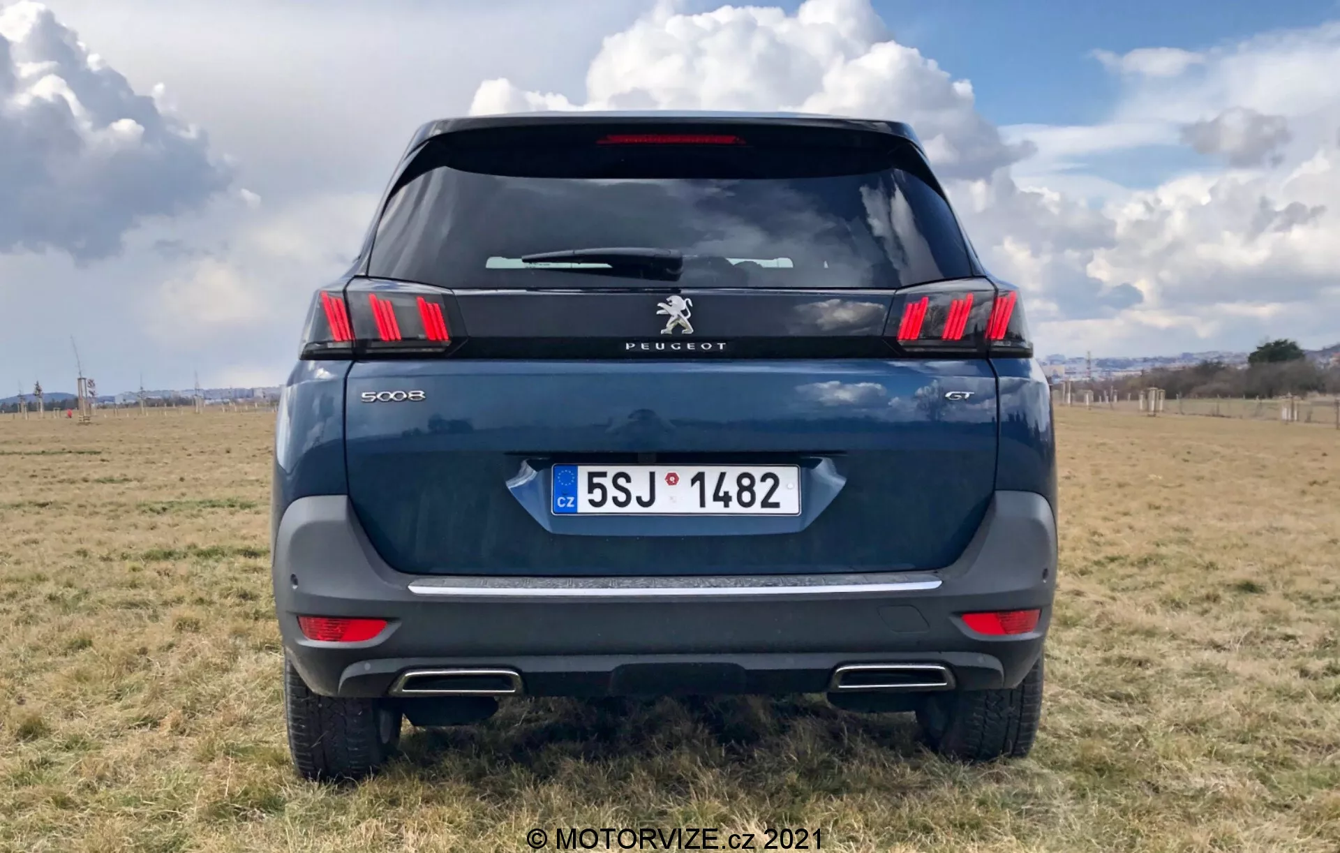 A modern Peugeot 5008 SUV hátsó nézete, kiemelve az elegáns vízszintes hátsó lámpákat jellegzetes karom alakú LED szegmensekkel, Peugeot logóval és a '5008' modell felirattal. Sötétkék vagy szürke színű, szabadtéri terepen parkol, részlegesen felhős ég alatt. A látható elemek közé tartozik a hátsó szélvédő, hátsó ablaktörlő, magasan felszerelt féklámpa, csomagtér fogantyú és a Cseh Köztársaság rendszámtáblája 'CZ' felirattal. A lökhárító tetején található a fényvisszaverő szalag, parkolószenzorokkal és a kipufogó végek kivágásaival. Az autó kitölti a kép nagy részét.