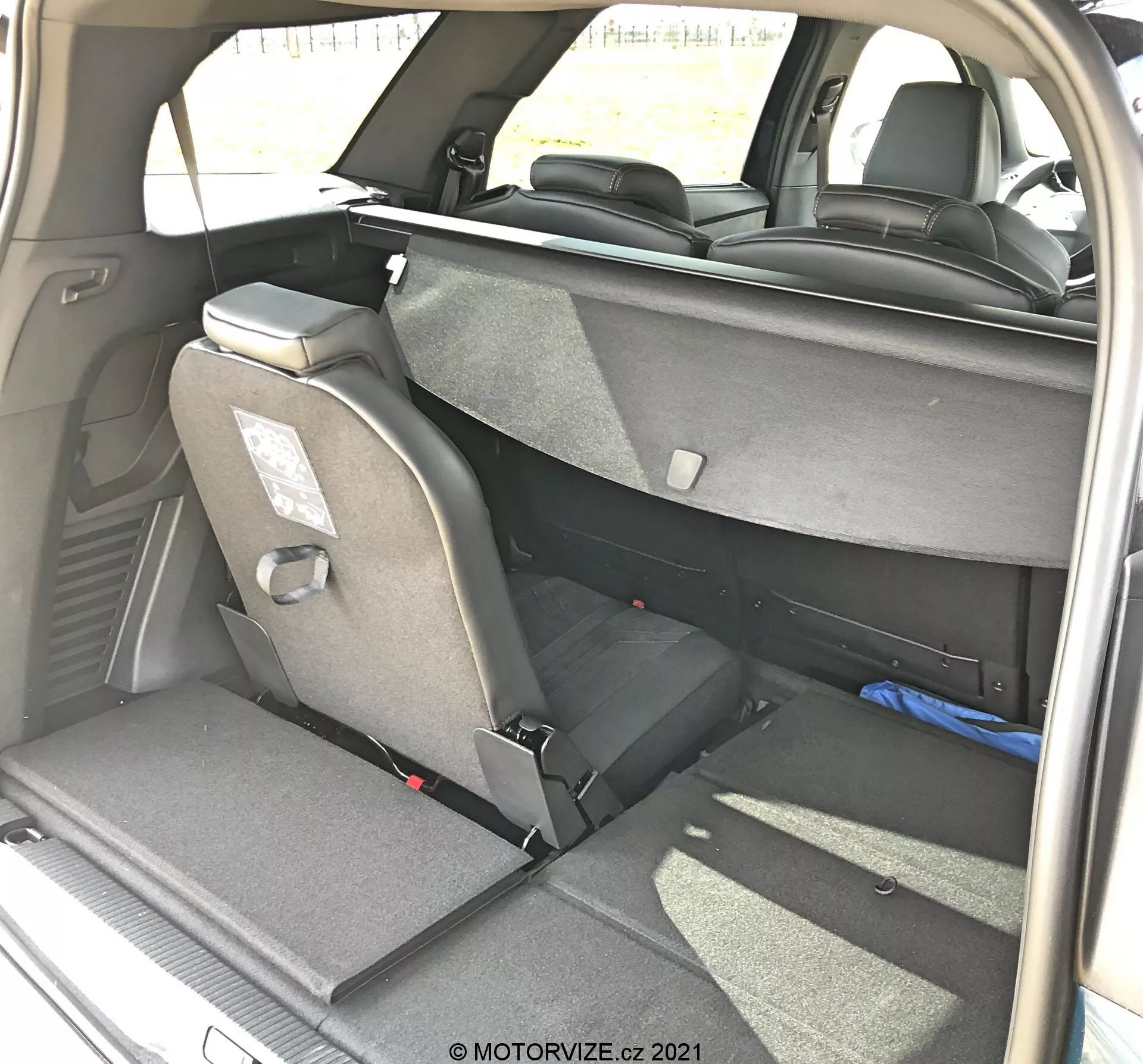 A Peugeot 5008 2021-es modelljének csomagtérbelső nézete jobb hátsó háromnegyedes szögből, rugalmas üléselrendezéssel, részben lehajtott hátsó ülésekkel, textil kárpitozással, fejtámlával és biztonsági övekkel. A jobb oldali hátsó ülés teljesen lehajtva, a bal oldali függőleges helyzetben maradva, jelezve a 60/40 osztható lehajthatósági funkciót. Látható a sötét belső kárpitozás és szőnyeg, természetes nappali világítással, motorvize.cz által készített felvétel.