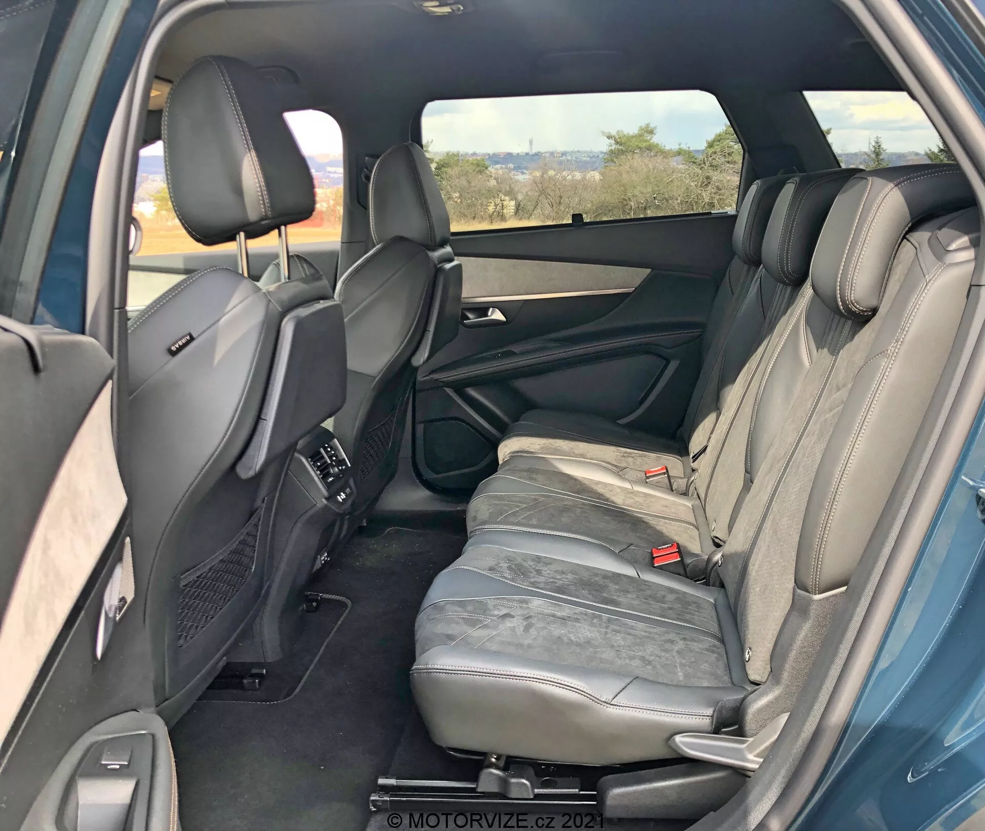 Egy Peugeot 5008 2021 belső tere a jobb hátsó nézőpontból, amely a hátsó utas teret mutatja. A jobb hátsó ajtó nyitva van, feltárva az ajtópanel jellemzőit, például a fogantyút, tárolórekeszeket és a hangszórót. Három szürke bőrrel kárpitozott hátsó ülés fejtámlával látható. A középső ülés kartámlája függőleges helyzetben van. A hátsó biztonsági övek és az állítható fejtámlával ellátott első ülések, valamint a középkonzol is látható. A középkonzolon elhelyezett hátsó klímaszabályozó szellőzőnyílások, színezett jobb ablak, a bal hátsó ajtó szimmetriáját jelző részletek. Összességében jól karbantartott belső tér látható technológiai funkciók nélkül.