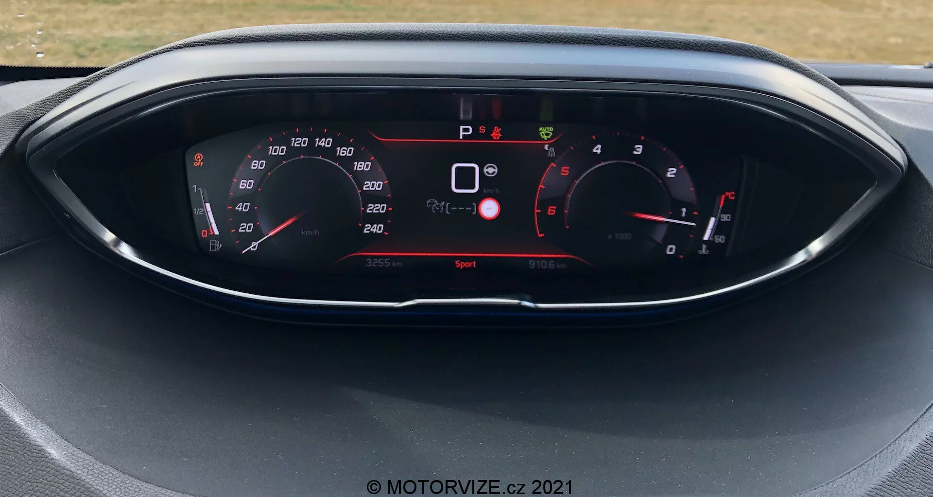 Közeli kép a 2021-es Peugeot 5008 műszercsoportjáról, digitális kijelzővel és analóg stílusú műszerekkel. A kijelző mutatja a sebességmérőt, fordulatszámmérőt, sebességfokozat kijelzőt és különböző jármű jelzőket, például biztonsági figyelmeztetéseket és vezetési módot. Az autó áll, a 'Sport' vezetési mód aktív.