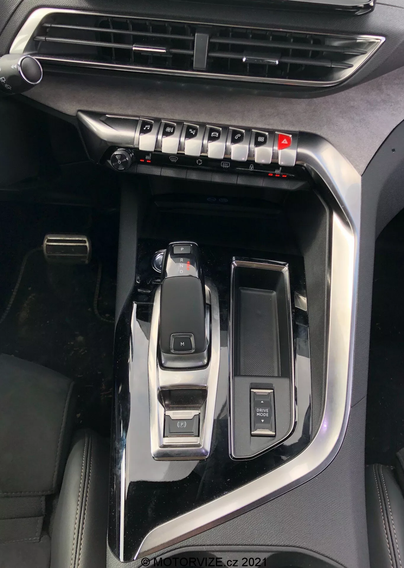 A Peugeot 5008 2021 középkonzolának közeli nézete, amely kiemeli a fontosabb jellemzőket, mint például a parkoló állásban lévő automata váltókar, az eco, sport, comfort vezetési módok közötti váltáshoz használt vezetéssel kiválasztó kapcsoló, a kulcs nélküli indításhoz használt start/stop gomb, a multimédia vezérlő forgókapcsoló, a jól látható légbeömlő az éghajlat szabályozásához és a zongora fekete felület fémes díszítéssel, amelyek prémium modellre utalnak, a vízjel 'MOTORVIZE.cz 2022'.