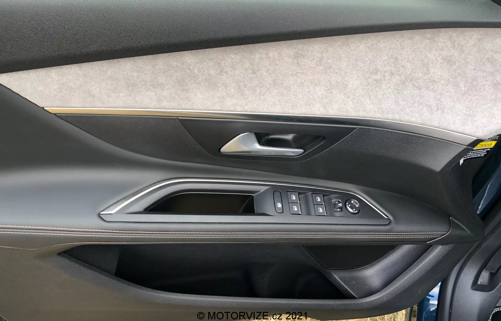 Közelkép a Peugeot 5008 2021 belső ajtópaneljéről, amely filc vagy szövet kárpitozású résszel, két tónusú formázott műanyag kialakítással rendelkezik, ahol a felső rész sötétebb, az alsó világosabb árnyalatú. Kiváló minőségű fémes vagy krómozott ajtókilincs, polírozott díszítőelemek és ergonomikus gombok találhatóak rajta az elektromos ablakemelőkhöz, az ajtózárhoz és az oldalsó tükrökhöz. Az ajtópanel alsó részén egy tárolózseb is látható.