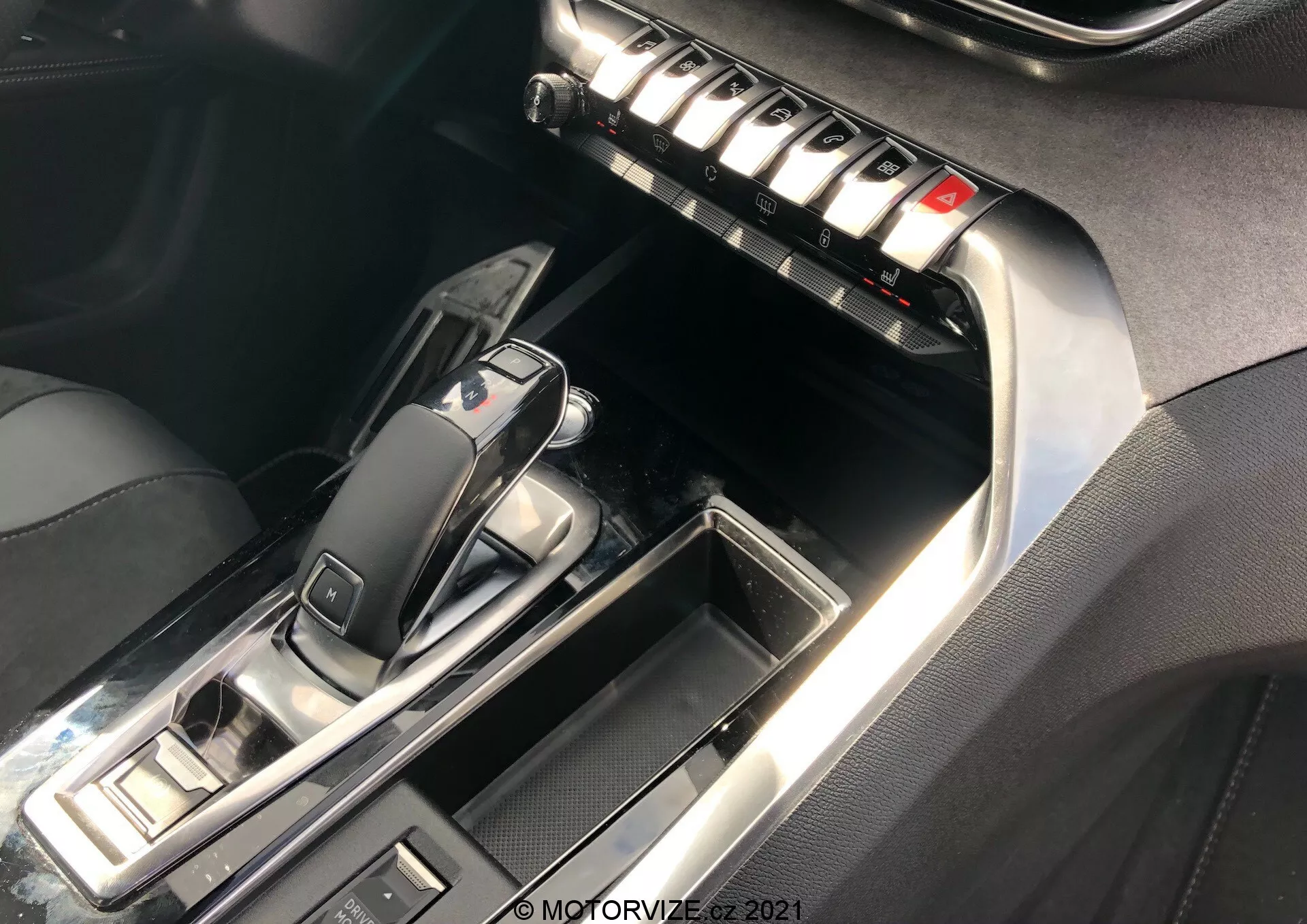 Közelkép a Peugeot 5008 2021 középkonzoljáról és első belteréről, különös tekintettel az automata váltóterületre és a központi egységre. A váltókar 'P' pozícióban van, modern, elegáns dizájnnal, fekete fényes műanyagból és fémes díszítéssel. A környező gombok vezetési módokra és rögzítőfékre utalnak. A középkonzolon egy sor fémből készült gomb található, valószínűleg a klímaberendezés, a páramentesítők és az ülések beállításaihoz. A kontrasztvarrás a bőrön vagy bőrhatású anyagon és a króm díszítések növelik a luxusérzetet. A világítás kiemeli a fényvisszaverő felületeket és a részleteket.