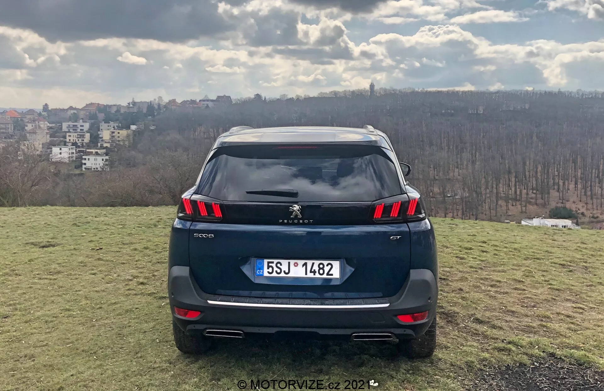 Egy Peugeot 5008 2021 SUV egyenes hátsó nézete emelkedett füves területen, városi háttérrel, amely LED-es hátsó lámpacsoportokat, a lökhárítóba integrált parkolószenzorokat, esetleges hátsó kamerát, európai stílusú rendszámot, kettős kipufogóvégeket, tetőspoiler és cápafarok antennát mutat; nappali jelenet, amely részleges felhőzetet sugall, a MotorVize.cz által 2022-ben fényképezve.