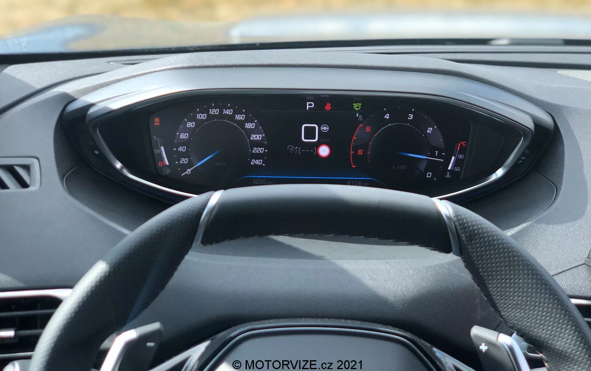 Modern jármű belső nézete első személy perspektívájából, a kormánykerékre és a digitális műszerfalra összpontosítva. A háromküllős kormánykerék középre helyezkedik el, mögötte világító műszerfal különféle műszerekkel és jelzőkkel. A sebességmérő és a fordulatszámmérő látható, km/h és mph értékekkel azonosítva, amely nemzetközi piacokra is alkalmas. A műszerfal számos figyelmeztető és tájékoztató fény jelent meg, mint például behúzott kézifék, nyitott ajtók, biztonsági öv figyelmeztetés és alacsony abroncsnyomás, fejlett fedélzeti diagnosztikát és biztonsági funkciókat jelezve. A műszerfal színsémája kék és piros, fekete háttéren fehér szöveggel. Ez a kép nappali fényben készült, és hangsúlyozza az autó belső kialakítását, a digitális műszerezést és a modern technológia.