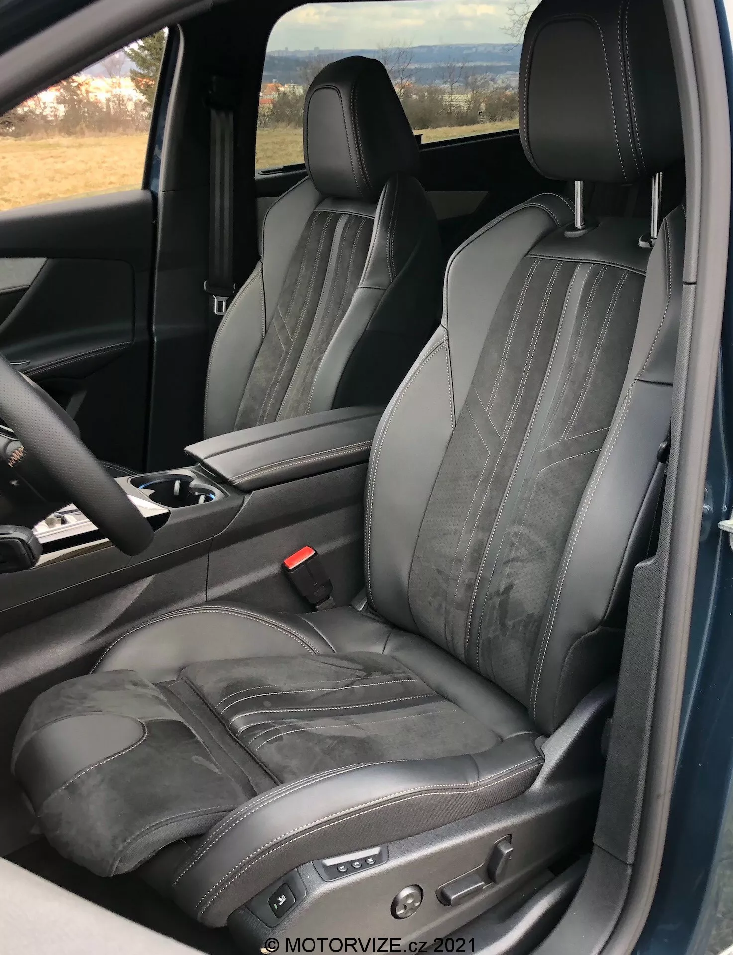 A Peugeot 5008 2021 belső nézete, amely a vezetőoldali ülésre összpontosít, állítható fekete bőrüléssel, kontrasztos varrással és vízszintes dombornyomással, nyitott ajtó perspektívájából készült, kiemelve a luxus és ergonomikus tervezést, elektronikus ülésbeállító vezérlőkkel és a középső konzol és a kormánykerék egy részével.