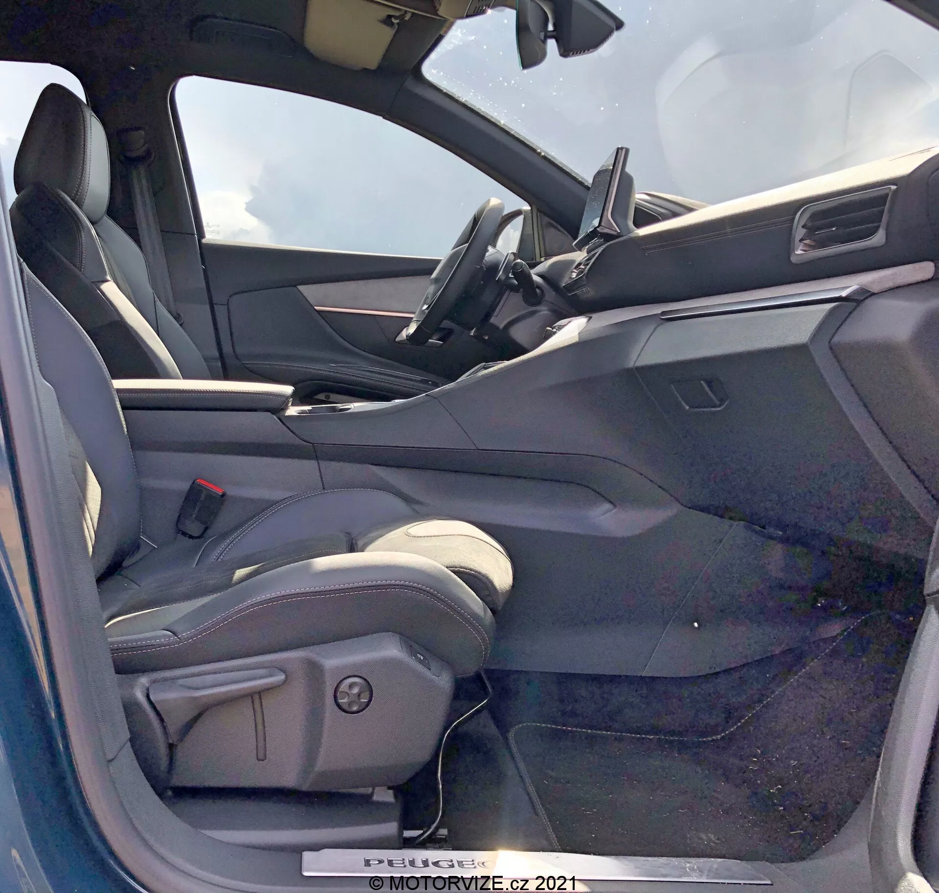 A 2021-es Peugeot 5008 belső nézete a sofőr oldaláról, nyitott ajtóval. A vezetőülés kagylós típusú, valószínűleg bőrrel vagy bőrhatású anyaggal kárpitozott, oldaltámaszokkal és elektromos állító vezérlőkkel. A nyitott vezetőoldali ajtón lévő belső panelen elektromos ablakvezérlők, valószínűleg elektromos tükörvezérlők és ajtózár mechanizmusok láthatóak. A műszerfal tartalmaz egy középre helyezett érintőképernyős infotainment kijelzőt, minimalist dizájnnal. A kormánykerék felső része látható, ami bőrrel bevont kormányt sugall beépített vezérlőkkel. A belső tér kéttónusú színvilágú, sötétebb felső felületekkel és világosabb alsó felületekkel.