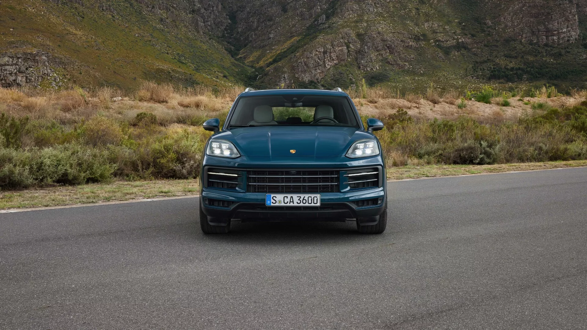 2023-as Porsche Cayenne (9YA Lifting) szemből, szemmagasságban látható, kiemelve az LED fényszórókat, markánsan megjelenő hűtőrácsot a márka logójával, sportos motorháztetőt és a beépített parkoló szenzorokat, hegyi háttérrel, ami luxust és kalandot sugall