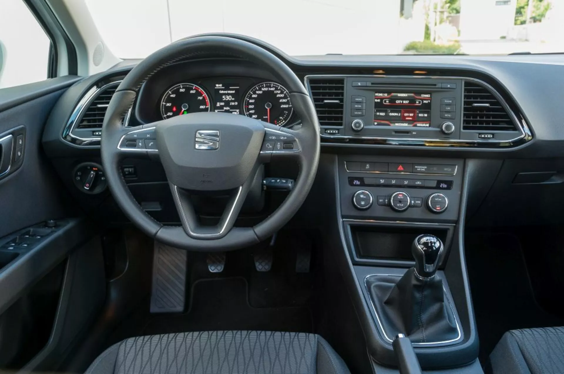 2016-os Seat Leon SC belső nézete az első utasülés perspektívájából, bemutatva a gyártó logójával és szerelt vezérlőkkel ellátott kormánykereket, digitális műszerfalat, infotainment rendszert, klímavezérlést, kézi váltót, és a vezető ajtóját elektromos opciókkal.