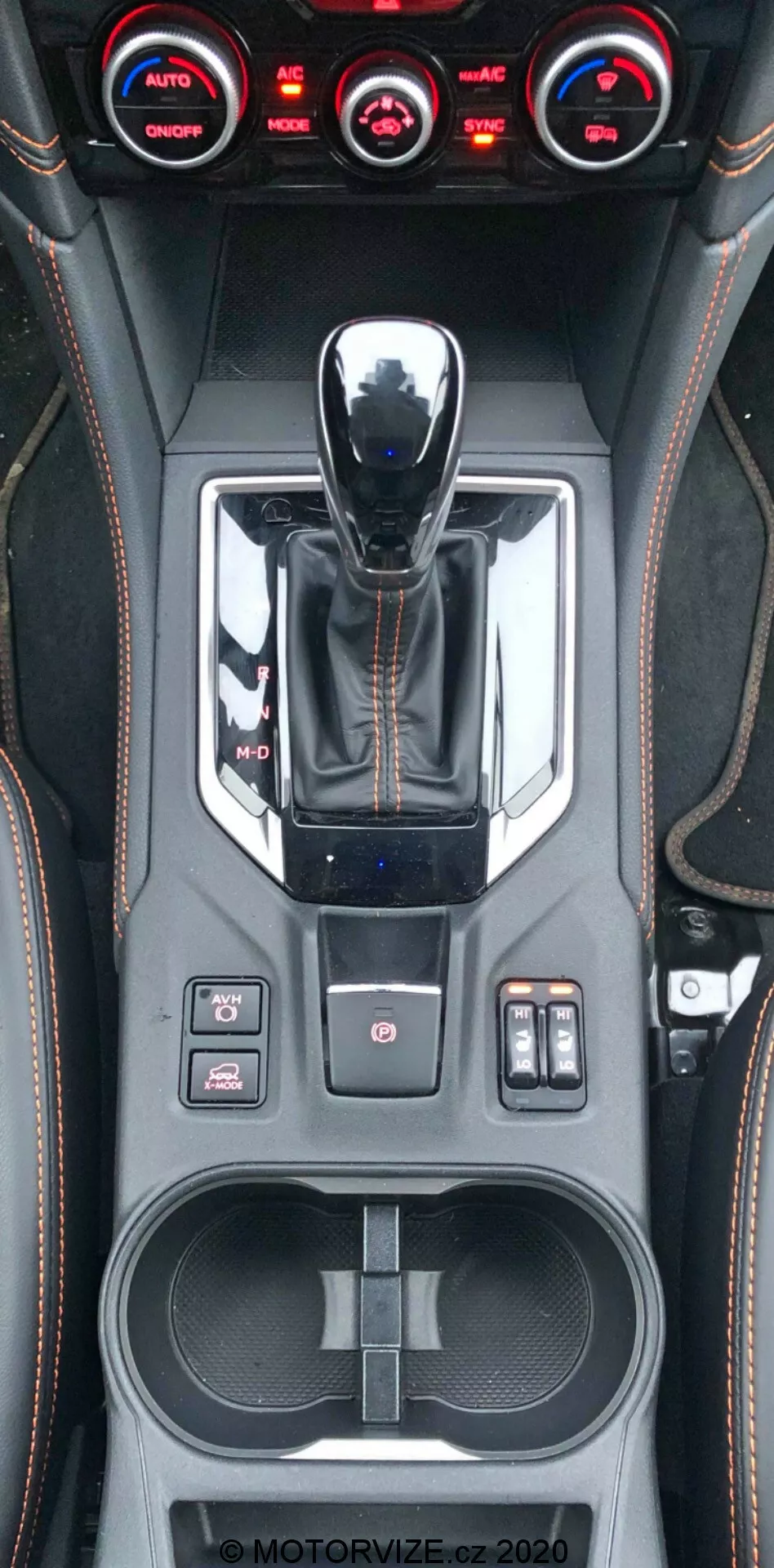 Függőleges közeli nézet a 2018-as Subaru XV középkonzoljáról, amely bemutatja a fémes kidolgozású váltókart, a vezetési mód választót, különféle vezérlőgombokat, beleértve a vonóerő-szabályozást, a parkolószenzorokat és a felfüggesztés beállításait, háromzónás klímaberendezést, kézzel varrott bőr-szerű díszítéseket, kéttálcás pohártartót, tárolóhelyet USB-portokkal, és látható MOTORVIZE.cz vízjelet.