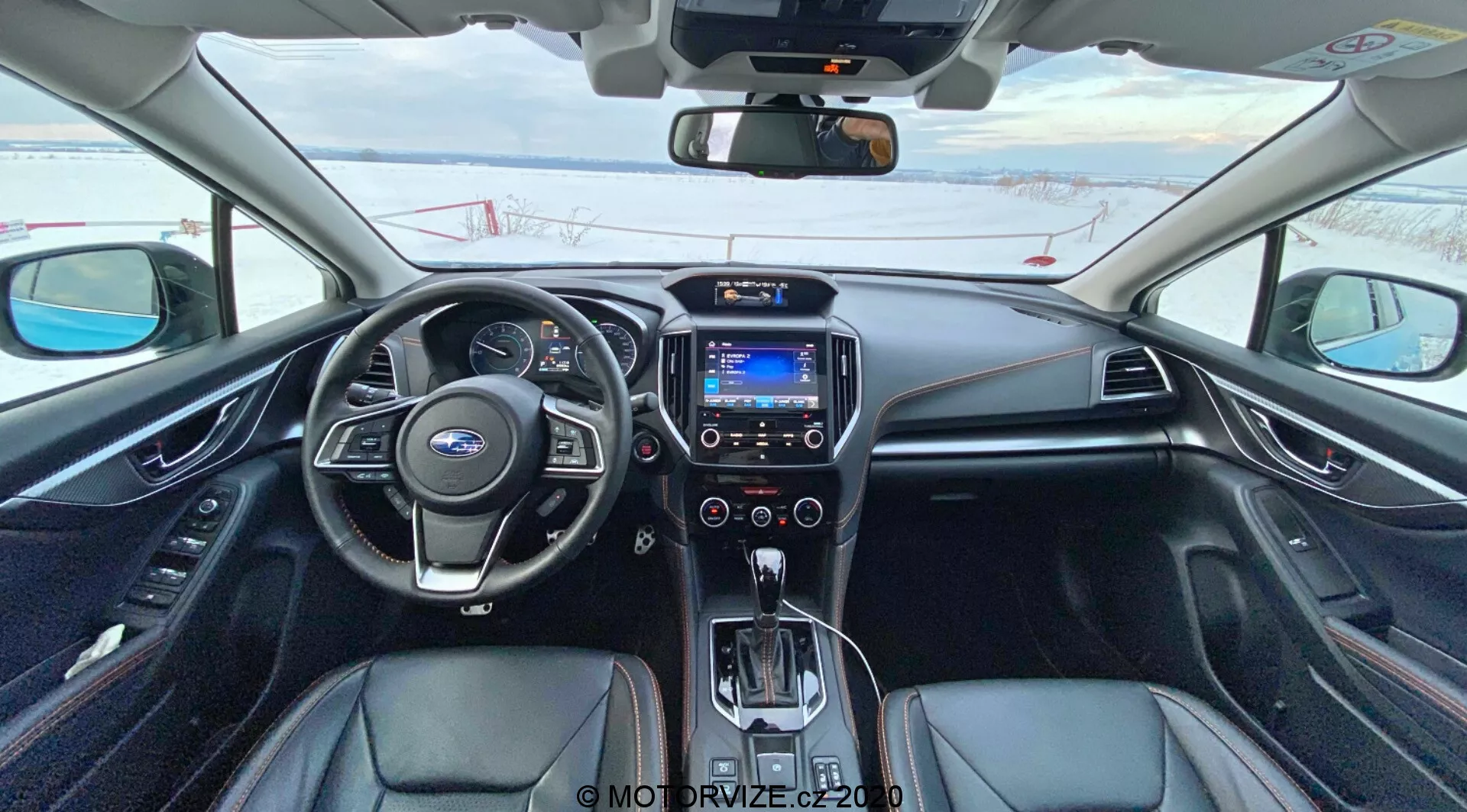 A 2018-as Subaru XV (Subaru XV 1.6i Trend Lineartronic) belső nézete a vezetőülésből, bemutatva a bőrbevonatú kormányt beépített irányítógombokkal, részben látható analóg és digitális műszercsoporttal, központi infotainment képernyővel kék interfésszel, klímavezérlő gombokkal, automatikus sebességváltóval, amelyet kezelőgombok vesznek körül, fekete kárpitozással ezüst díszítéssel, ülésfűtés vezérlőgombjaival és digitális kijelzővel rendelkező visszapillantó tükörrel. Az autó parkol, és odakint hó látható, ami hideg éghajlatot jelez.