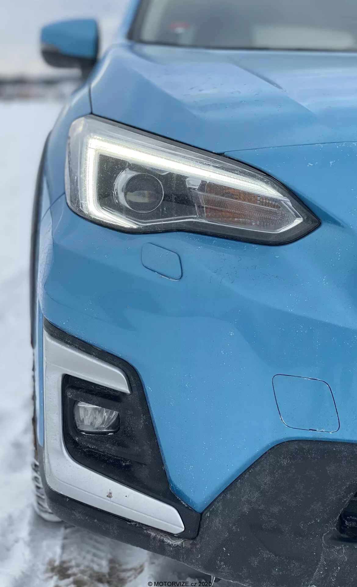 A 2018-as kék Subaru XV 1.6i Trend Lineartronic bal első oldalának közeli képe, ferde szögből készítve, amely a fényszóróra és a hűtőrácsra, valamint az első lökhárító egy részére fókuszál. A kiemelkedő bal oldali fényszóró vetítőlencsével rendelkezik, ami a HID vagy LED rendszereket jelzi, valamint integrált LED nappali menetfényekkel van ellátva. A kép az első lökhárító egy részét is mutatja, amely valószínűleg további világítást tartalmaz. Az autó karosszériája éles vonalakkal és aerodinamikus formákkal rendelkezik, téli háttérrel és enyhe hóréteggel.