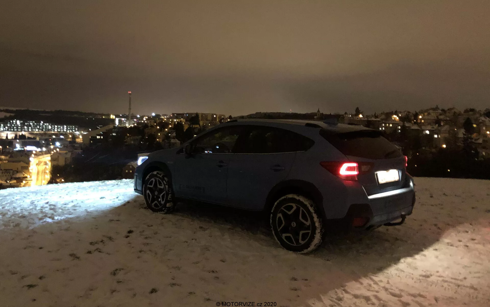 Subaru XV 1.6i Trend Lineartronic 2018 éjszaka hátulról háromnegyed nézetből fotózva, bemutatva a jobb oldalt és a hátulját, beleértve a hátsó lámpákat, hátsó lökhárítót, jobb hátsó kereket, utas ajtókat és a jobb első kereket. A jármű, valószínűleg sötétkék vagy szürke színű, havas felületen parkol, a távolban látható városi fényekkel. A hátsó lámpák arra utalnak, hogy az autó be lehet kapcsolva, és a jellegzetes, nagyobb kerekek egy magasabb felszereltségi szintet vagy sportos változatot jeleznek. A jelenet az esztétikai és környezeti elemeket hangsúlyozza anélkül, hogy az előrehaladott technológiai funkciók jól láthatóvá válnának.