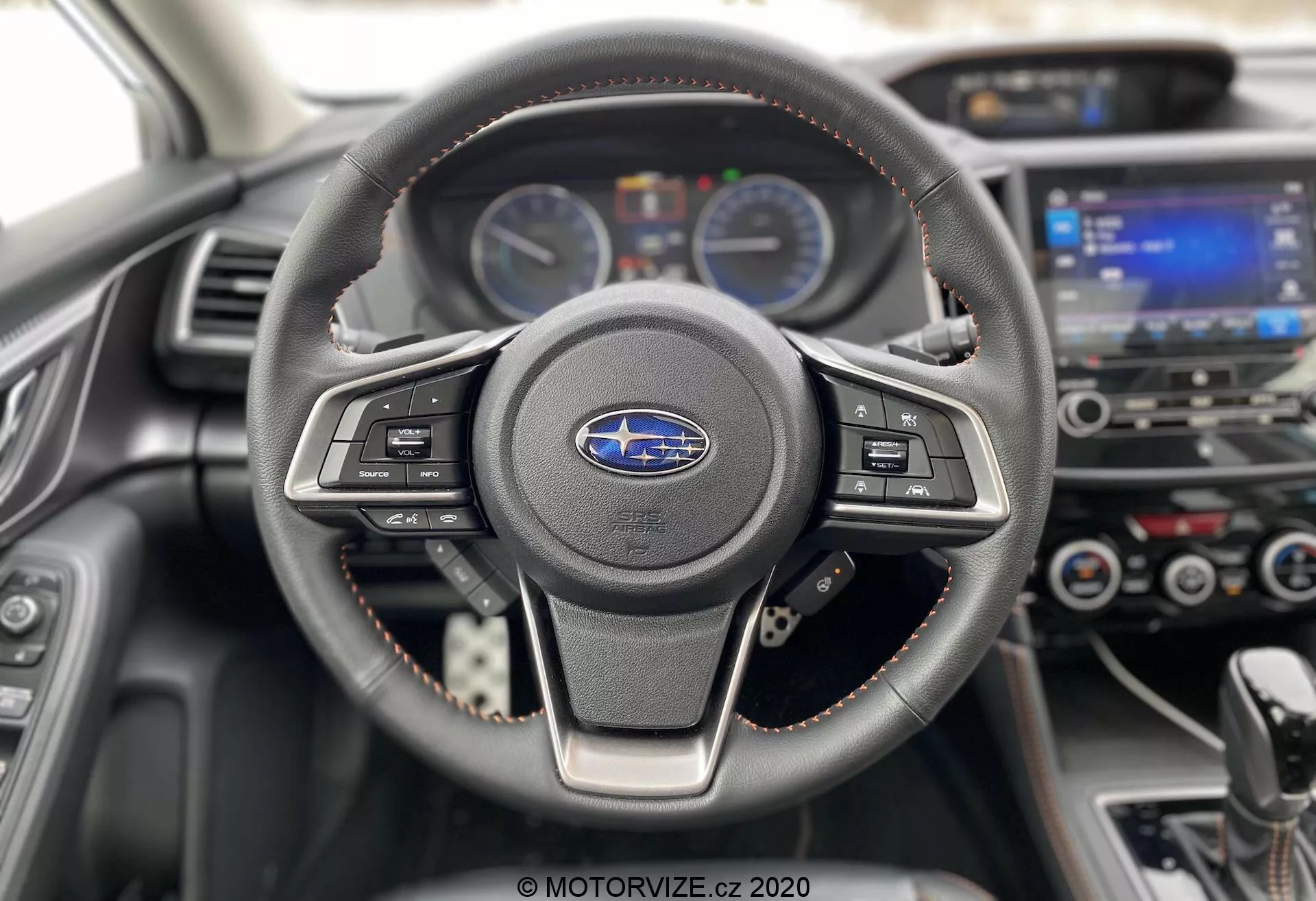 A Subaru XV 2018 (Subaru-xv-1-6i-trend-lineartronic) belső nézete, amely a kormányra és a műszerfalra összpontosít. A háromküllős, bőrből készült kormánykerék látható varrással rendelkezik, és multimédia és sebességtartó automatika vezérlőgombokkal van felszerelve. Mögötte a műszerfal két analóg mérőórát mutat, amelyeket egy digitális sofőrinformációs kijelző választ el. A háttérben, enyhén homályosan, érintőképernyős infotainment rendszer és klímavezérlők láthatók. A kormánykeréken és a műszerfal párnázatán lévő piros varrás kiemeli a sportos vagy prémium felszereltséget. A vezetőülés perspektívájából készült, modern és jól karbantartott belső kép hangsúlyozza az autótechnológiát és a dizájnt.