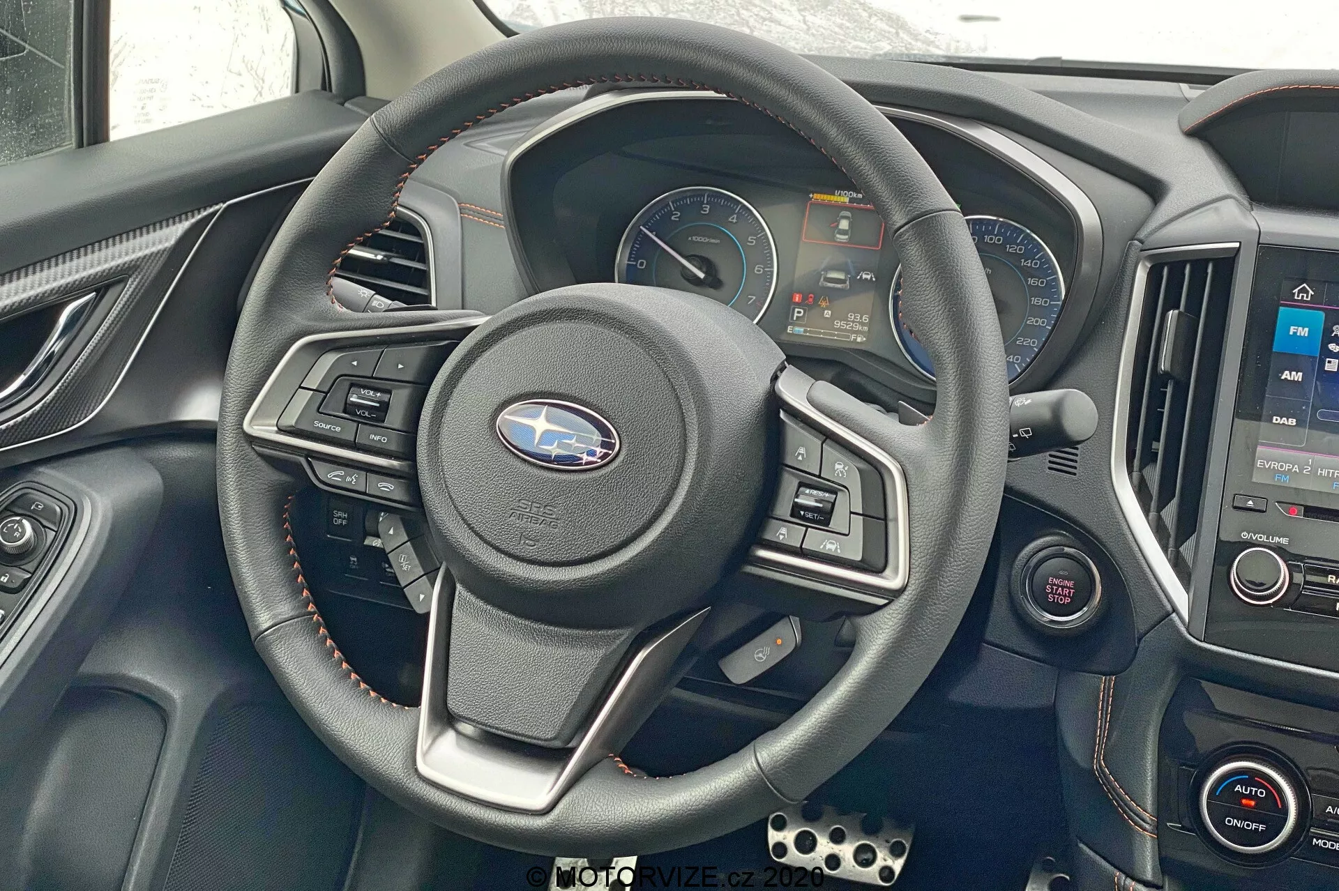 Subaru XV 2018 belső felvétel, a kormánykerékre és a műszerfalra összpontosítva. Középen elhelyezett háromküllős kormánykerék beépített vezérlőkkel, digitális műszercsoport sebességmérővel, fordulatszámmérővel és információs kijelzőkkel. Középkonzol infotainment rendszerrel, amely térképet jelenít meg, alatta klímavezérlők. Fémes díszítések, bőrrel burkolt kormánykerék, piros szellőzőrészletek. Bal oldali további vezérlőgombok, sportpedálok láthatók a bal alsó sarokban. Frontális, enyhén lefelé néző szög a vezetői kezelőszervek és a technológia bemutatására.