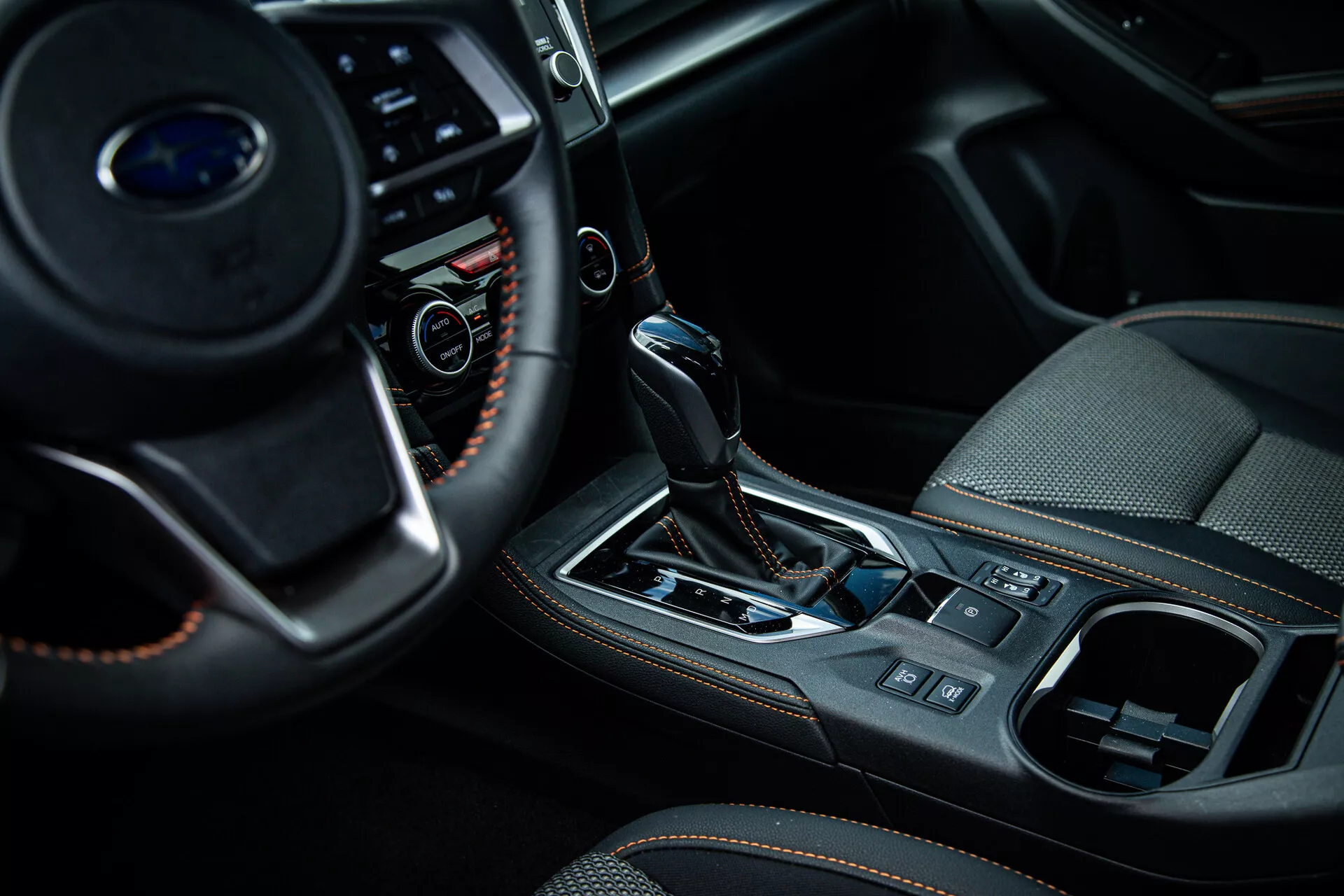 Közeli kép egy 2021-es Subaru XV belső teréről, a középkonzolra és a sebességváltó területére összpontosítva az utas oldalról nézve, bemutatva a kormányon lévő multifunkciós gombokat, az automatikus váltókar bőr burkolatával és fémes díszítőelemekkel, valamint a felső kategóriás belső kialakítás jellemzőit, mint az elektronikus kézifék és a klímavezérlő rendszer.