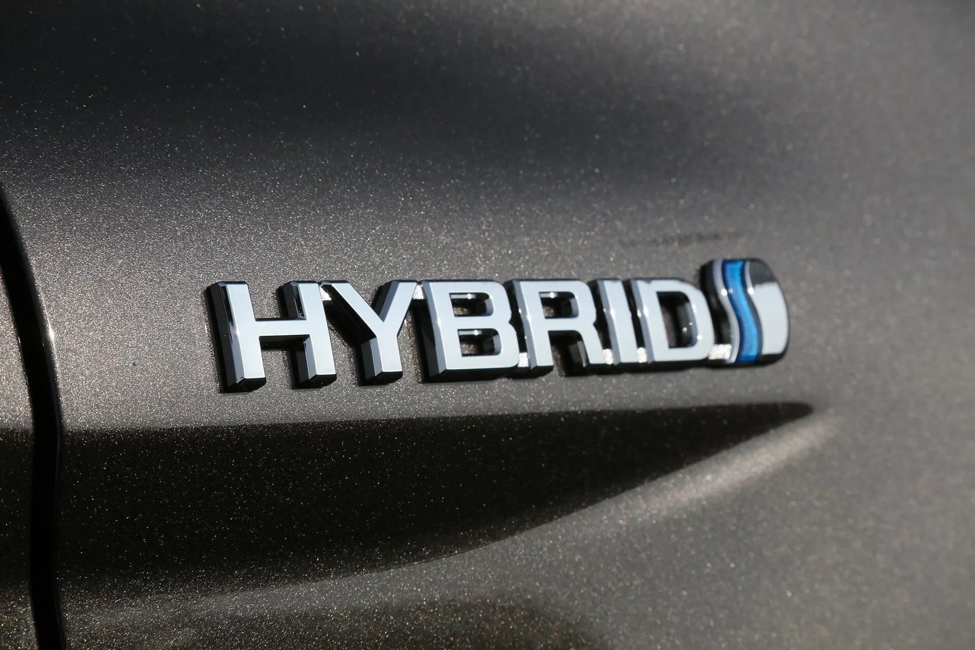 Közeli kép a Toyota Camry 2001 hibrid emblémájáról króm és kék kiemelésekkel, bemutatva az autó karosszériáján az ökobarát hibrid technológiát természetes fényben.