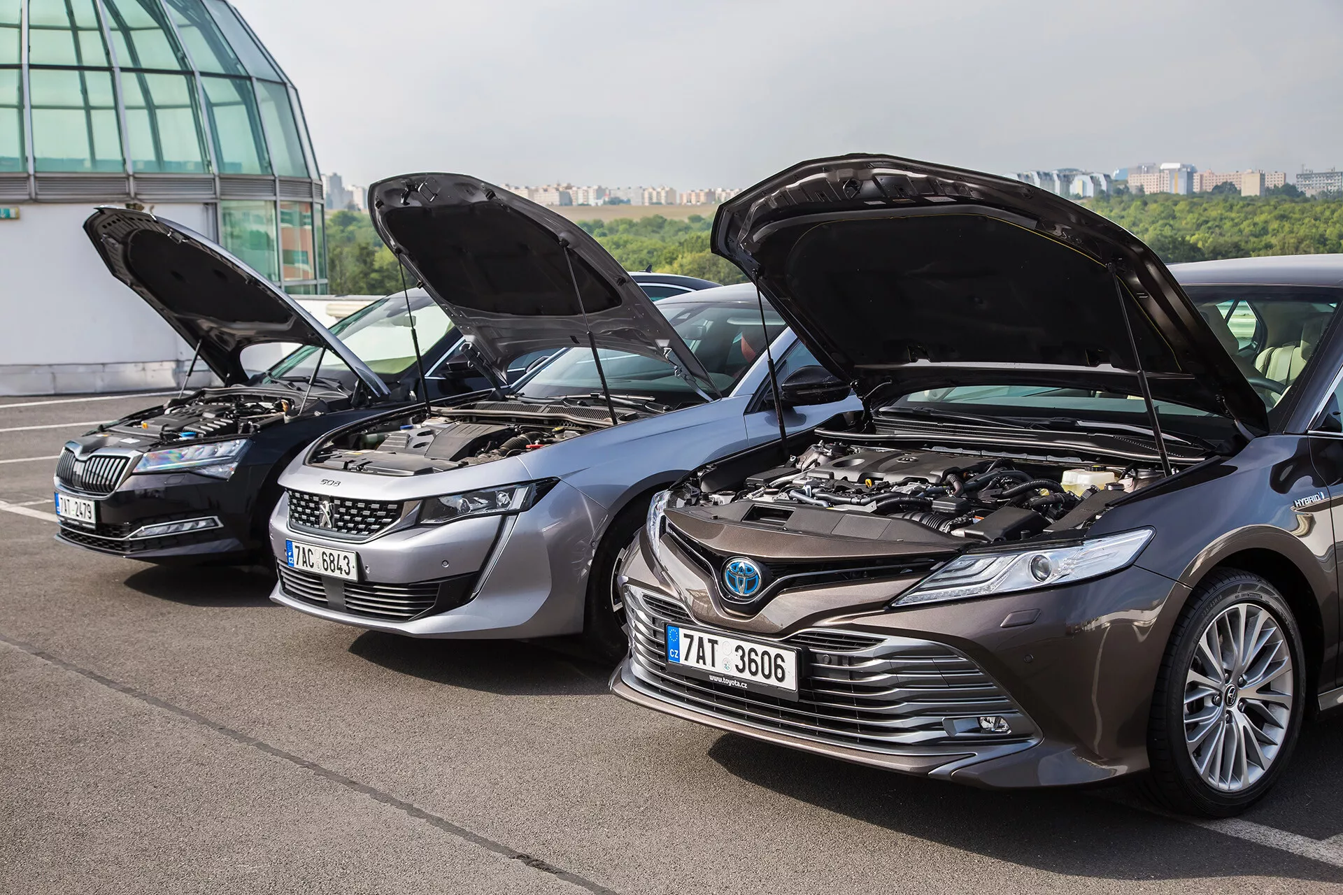 Toyota Camry 2001 bemutatva nyitott motorháztetővel a Skoda és a Land Rover mellett, feltárva a motorokat és belső alkatrészeit európai kültéri környezetben, épület háttérrel