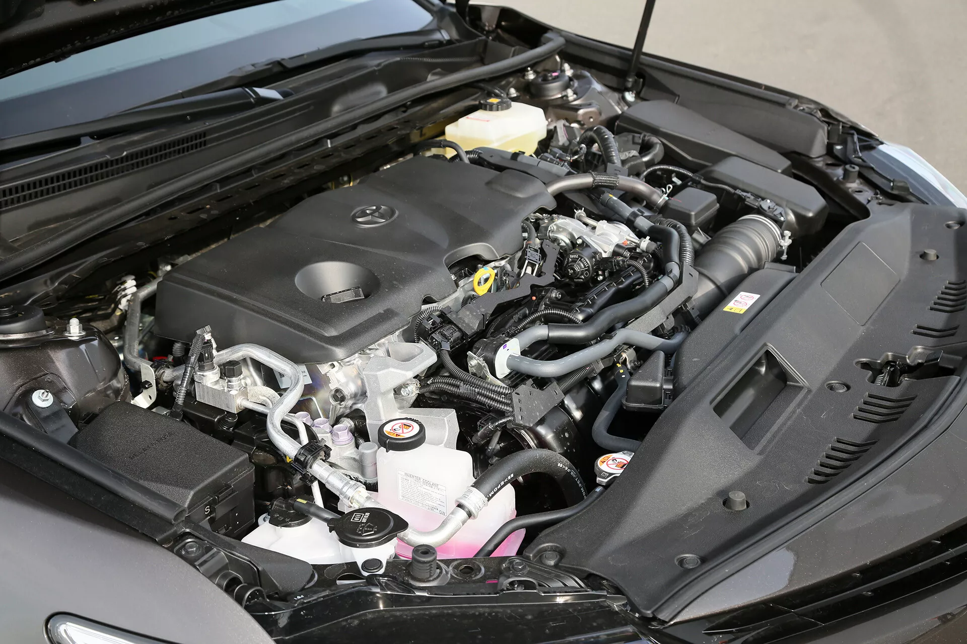 Közelkép egy 2001-es Toyota Camry limuzin nyitott motorházáról, kiemelve a motor fedelét a gyártó emblémájával, a levegőbeviteli rendszert, a folyadéktartályokat, az akkumulátort, a kábelezést, a csöveket, a biztosítékdobozt, a motor fém alkatrészeit és az első rész szerkezetét.