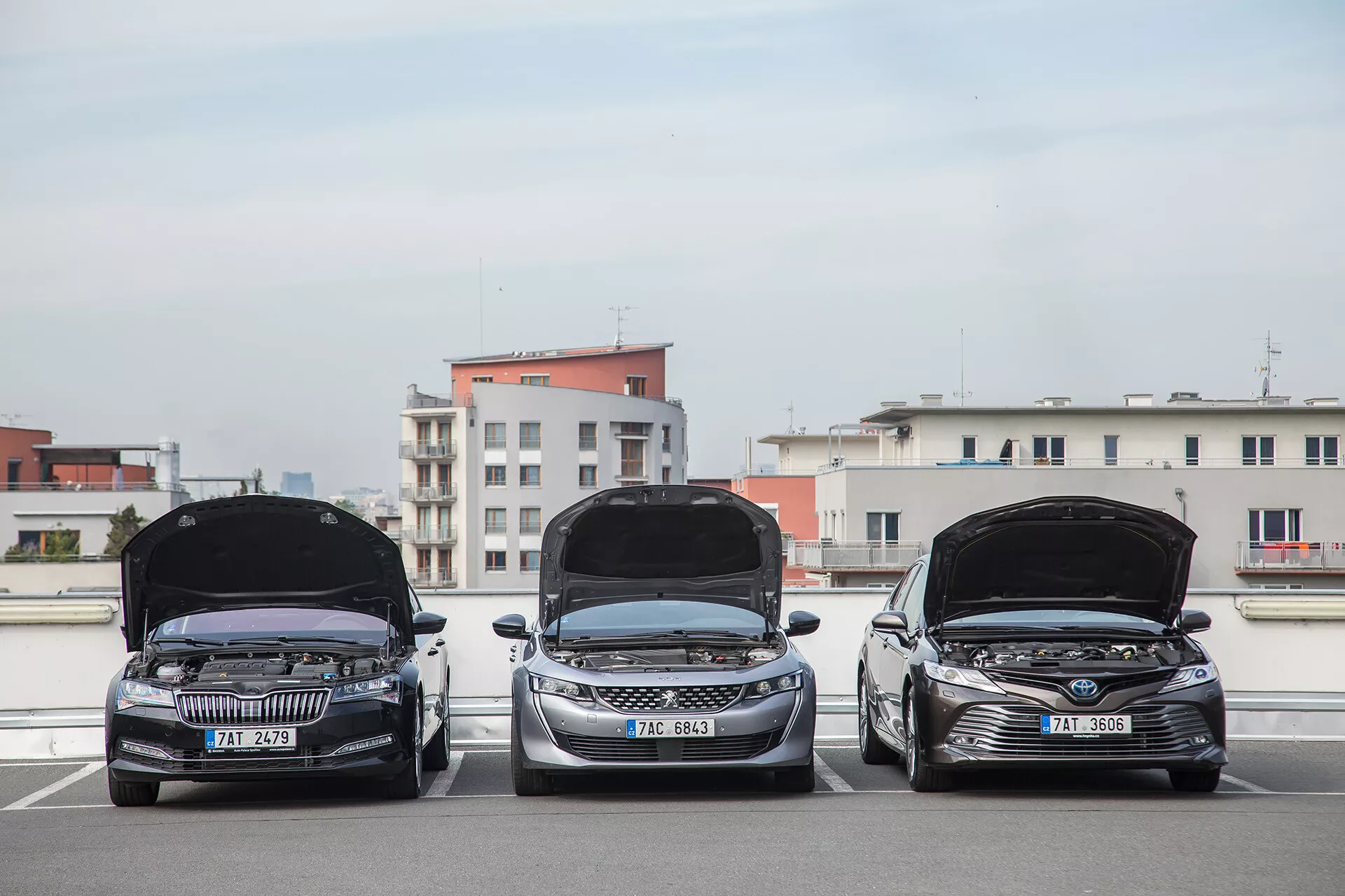 Három 2001-es Toyota Camry limuzin parkol egymás mellett egy tetőtéri parkolóban, az autók eleje és nyitott motorháztetők láthatóak, a fényes nappali fényben a technológiai jellemzők és a dizájn városi háttérrel