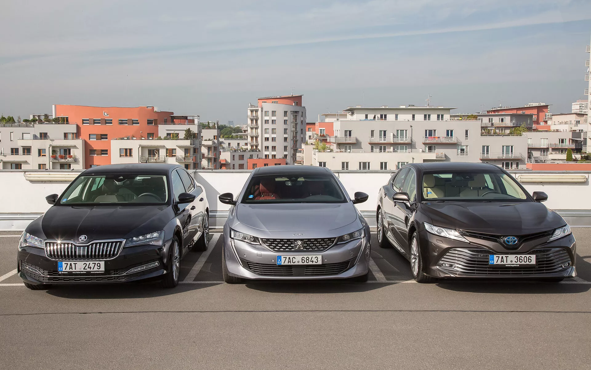 Skoda, Peugeot és Toyota szedánok oldalról való összehasonlítása háromnegyed előlnézetben egy tetőtéri parkolón, modern tervezési jellemzőket hangsúlyozva, mint az LED világítás, aerodinamikai formák és króm díszítőelemek