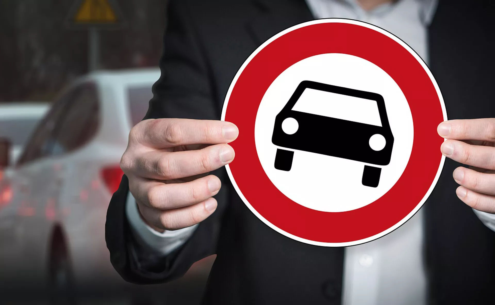 Egy személy tart egy piros kör alakú táblát fehér autó sziluett ikonnal, amely általános autó ábrázolást szimbolizál elmosódott városi háttérrel szemben